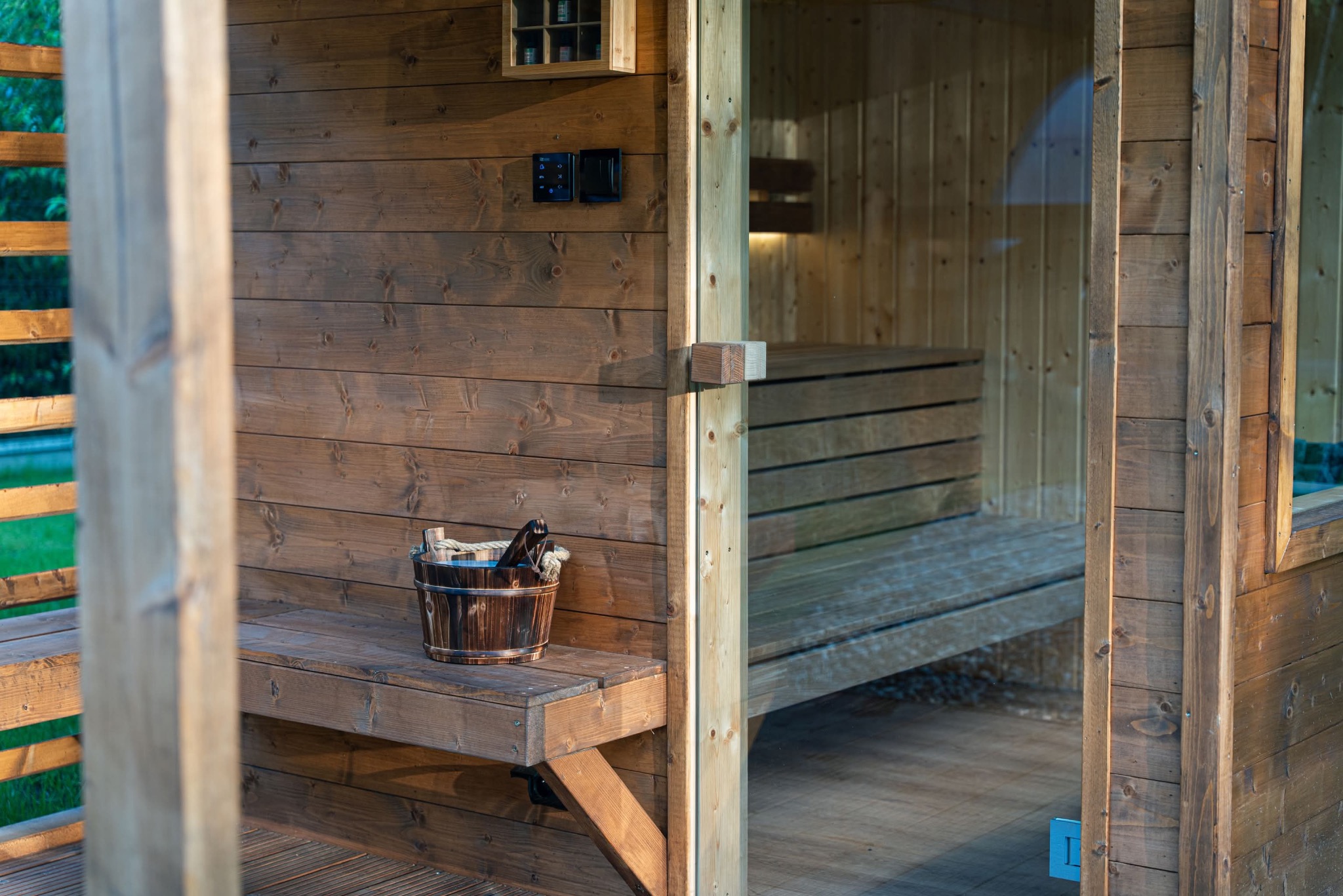 Drewniana sauna z przeszklonymi drzwiami, widoczne wnętrze z ławkami i wiadro na zewnątrz. Ciepłe oświetlenie, naturalne drewno, detale wykończenia.