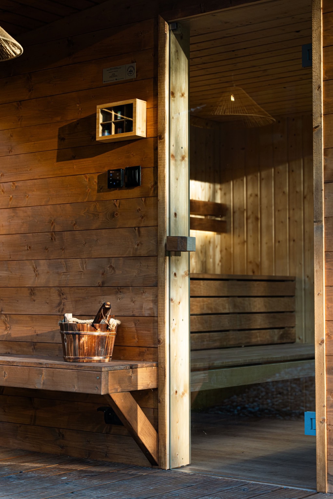 Drewniana sauna z przeszklonym wejściem. Wnętrze oświetlone, widoczne ławy. Na zewnątrz wiadro z akcesoriami na ławce. Ciepłe światło podkreśla naturalny charakter drewna.