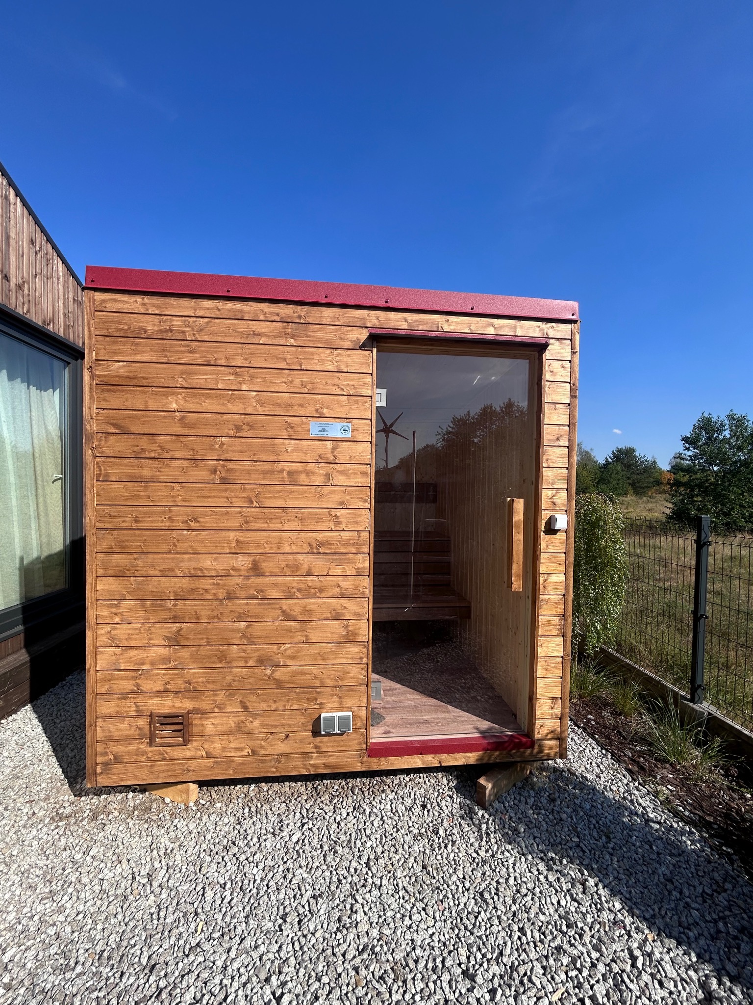 Gotowa, drewniana sauna zewnętrzna z przeszklonymi drzwiami, ustawiona na żwirowym podłożu w słoneczny dzień. Czerwone wykończenia dachu i drzwi.