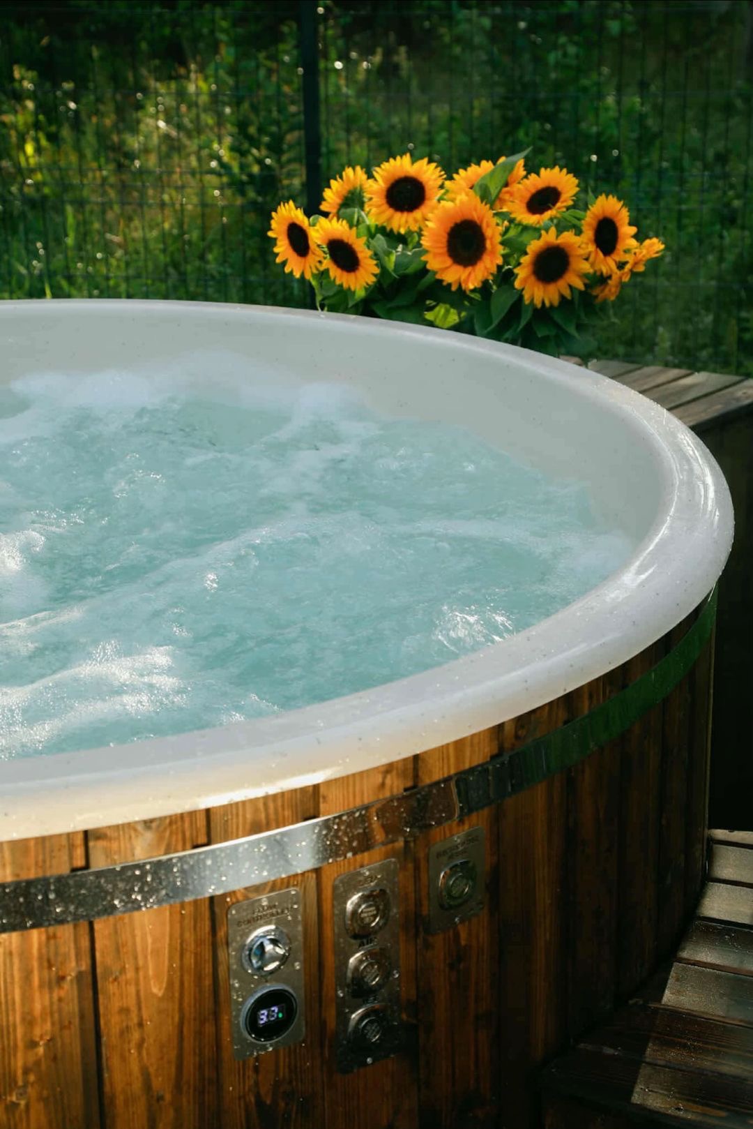 Drewniana balia ogrodowa z hydromasażem, wypełniona wodą. W tle zielona roślinność i słoneczniki w donicy. Widoczne sterowanie balii.