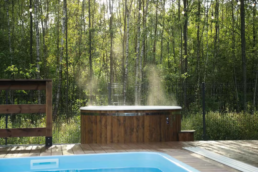 Drewniana sauna beczka na tarasie, otoczona lasem. Para wodna unosi się z sauny. Częściowo widoczny basen. Oświetlenie dekoracyjne. Słoneczny dzień.
