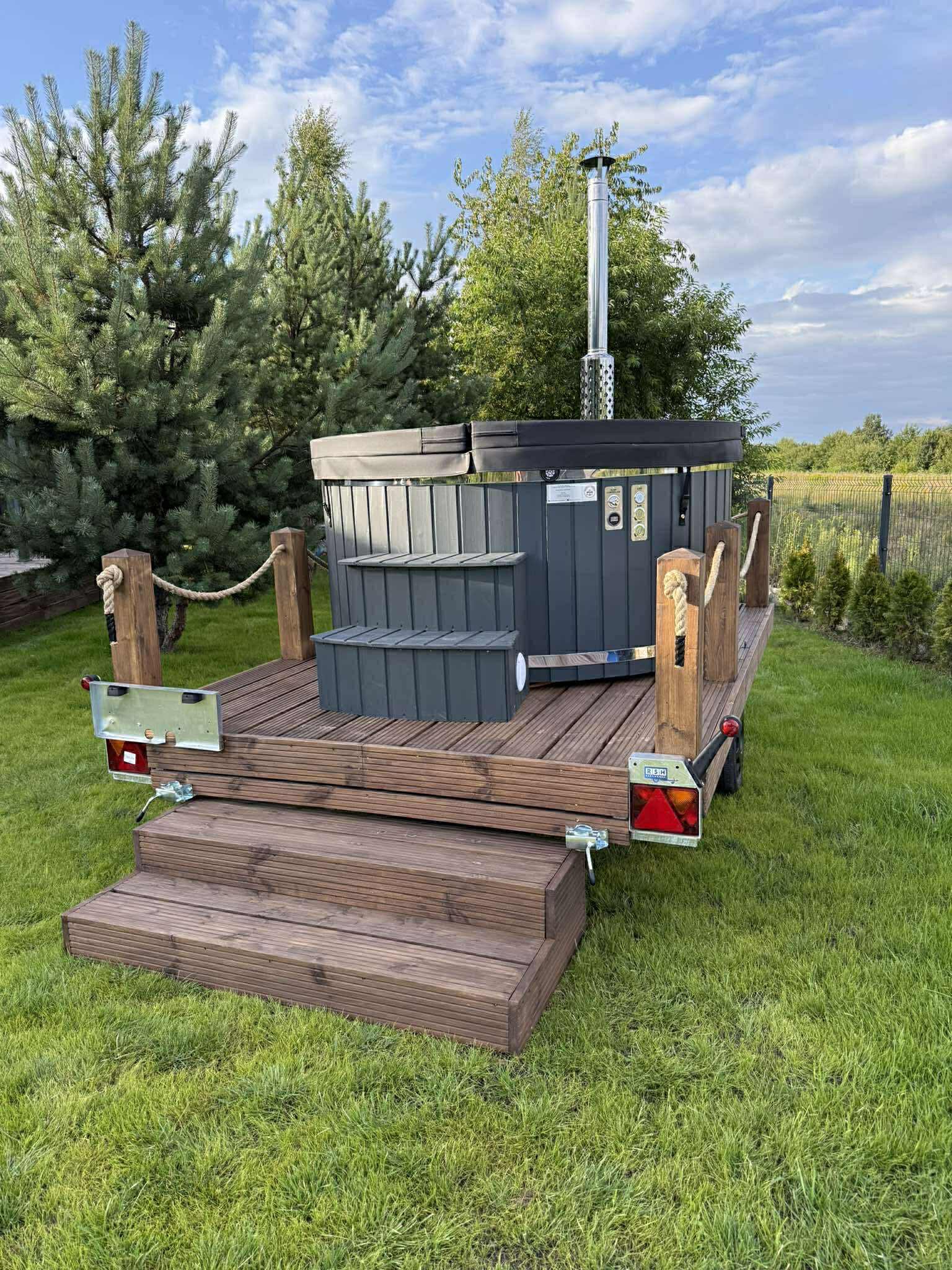 Mobilna sauna na drewnianym podeście z schodkami, umieszczona na przyczepie, otoczona zielenią. Widoczny komin sauny i elementy dekoracyjne z liny.