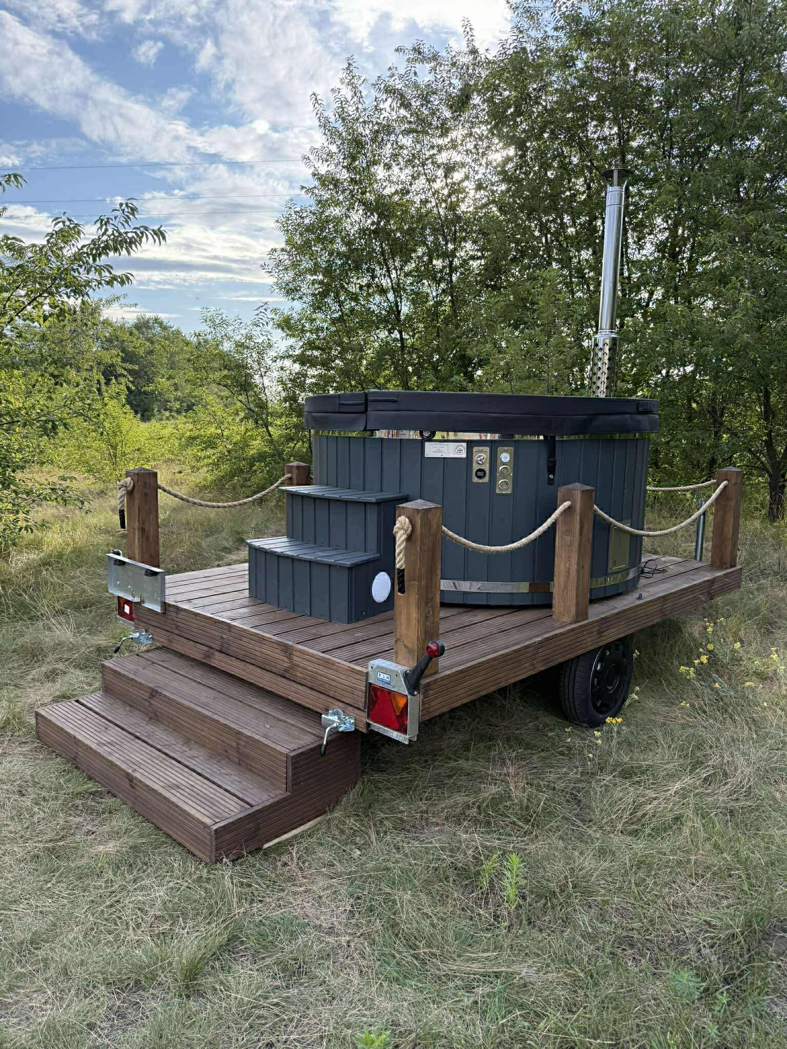 Przenośna sauna na drewnianym podeście z schodkami, umieszczona na przyczepie, otoczona zielenią. Kompaktowa konstrukcja z kominem i drewnianymi balustradami.