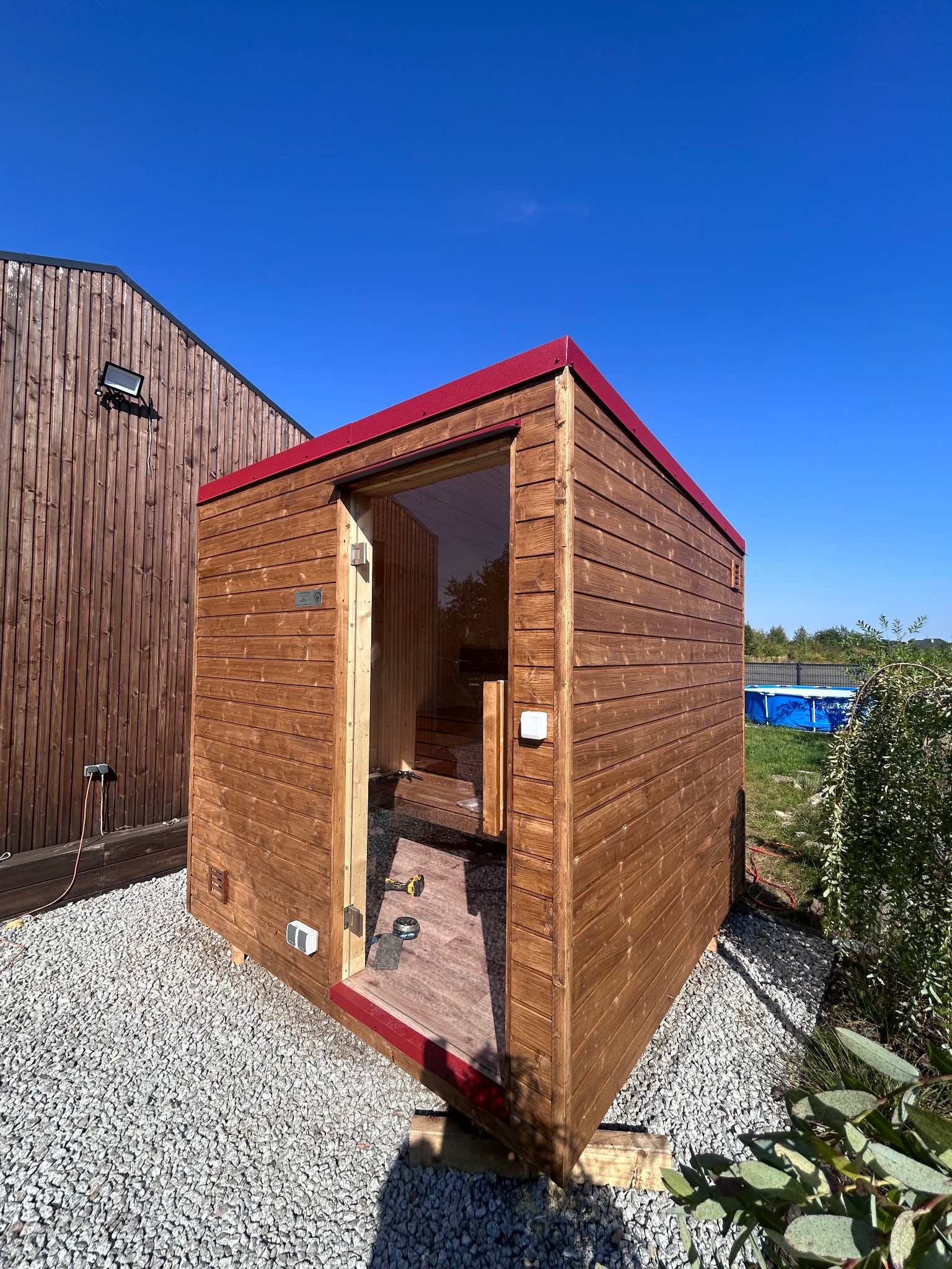 Nowoczesna, drewniana sauna zewnętrzna z czerwonymi akcentami, częściowo otwarta, ukazująca wnętrze i narzędzia. W tle błękitne niebo i budynek z drewnianą elewacją.