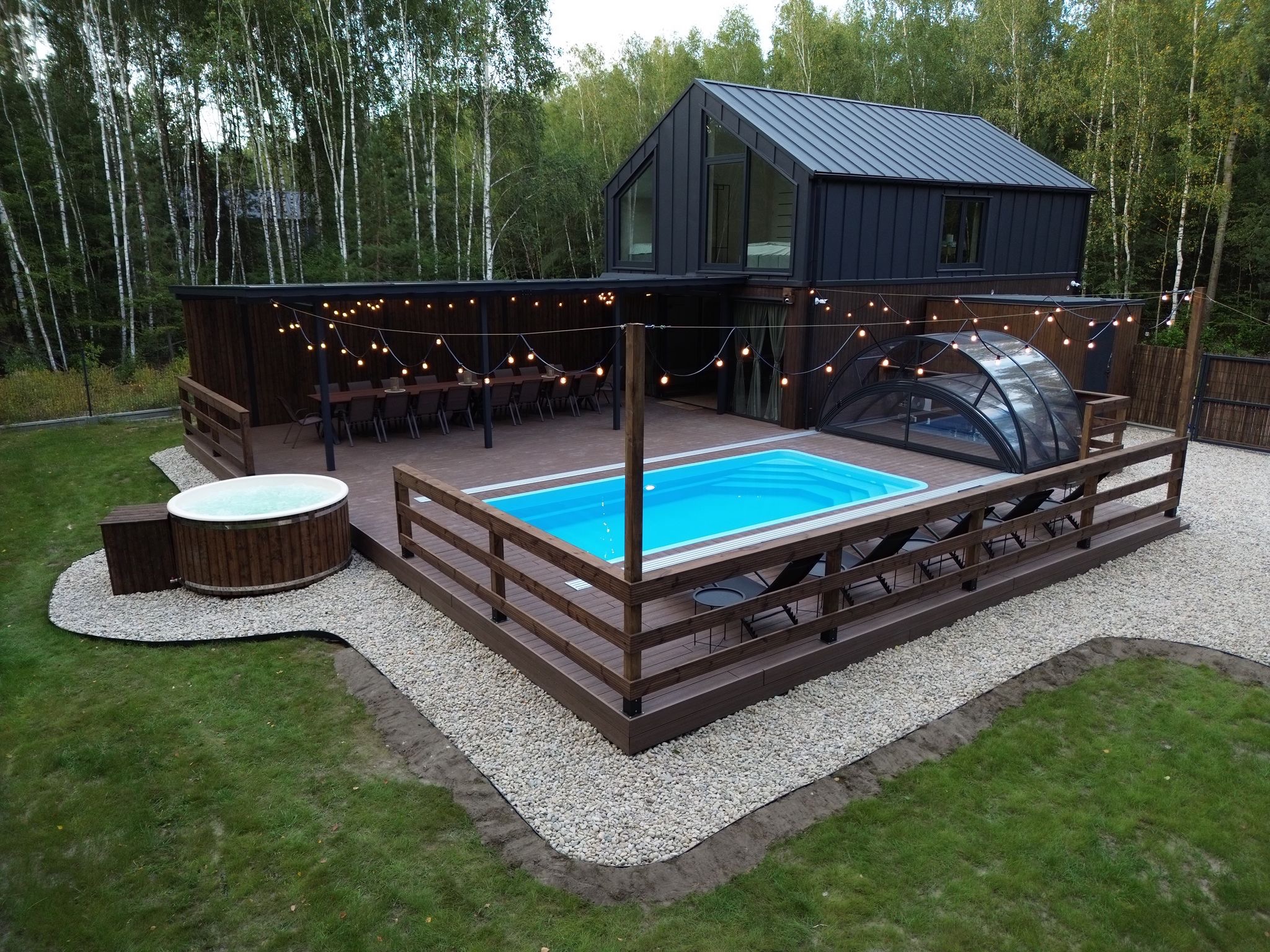 Nowoczesny taras z basenem i jacuzzi, otoczony drewnianą zabudową i dekoracyjnym oświetleniem. W tle dom z ciemną elewacją i las.