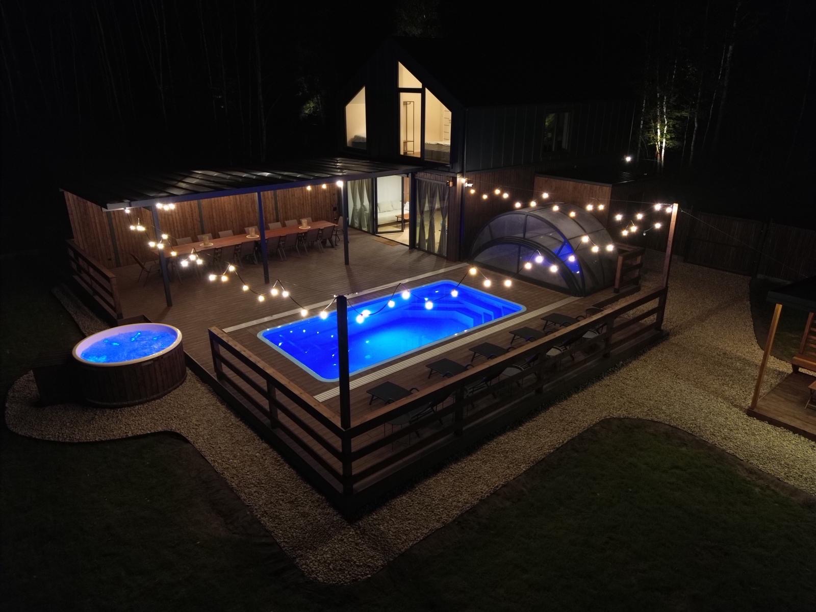 Oświetlony basen i jacuzzi na drewnianym tarasie nocą. W tle budynek z oknami i zadaszona jadalnia. Dekoracyjne lampki dodają uroku przestrzeni.