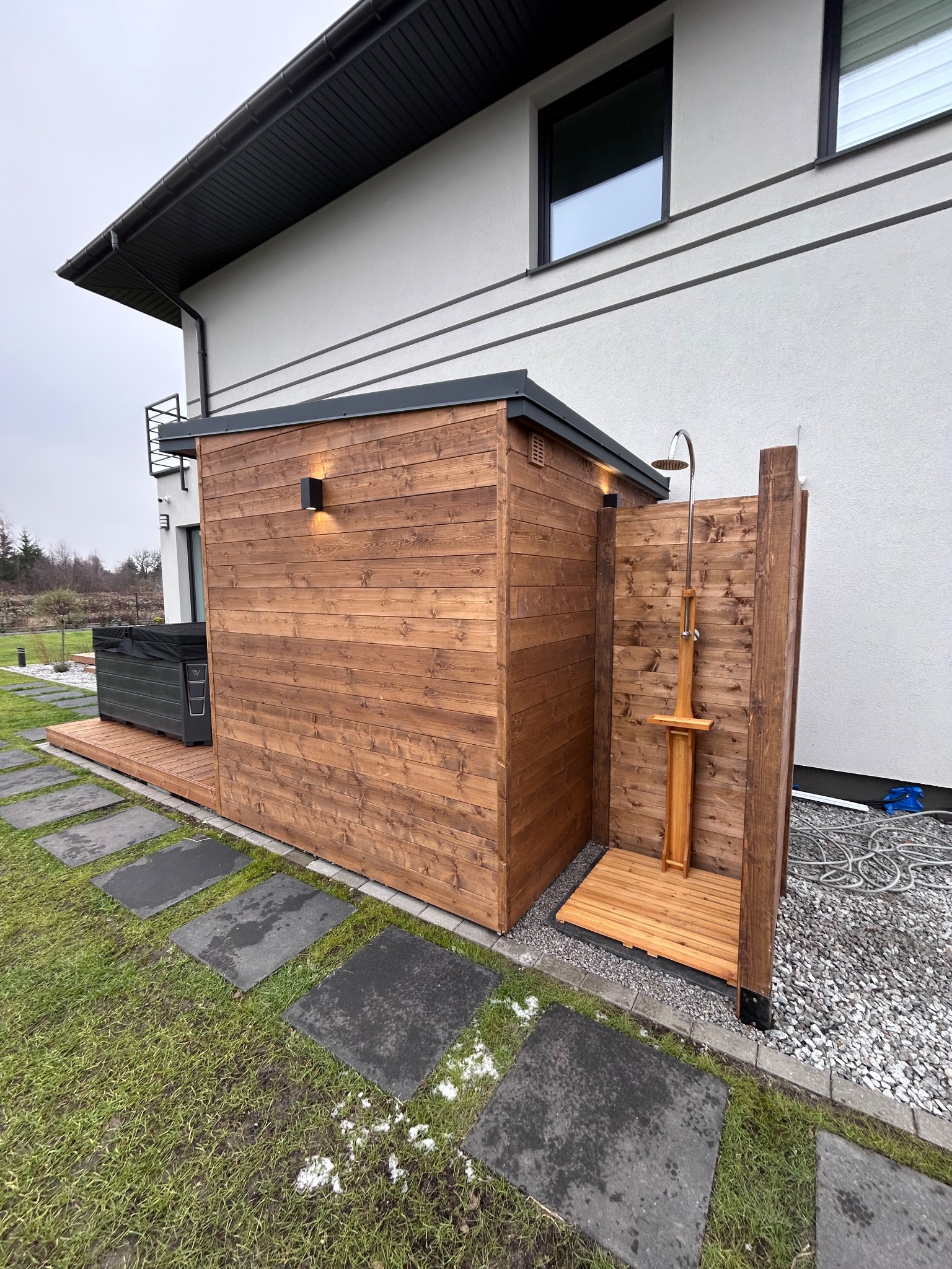 Drewniana sauna zewnętrzna z prysznicem, obok jacuzzi na tarasie. Całość zlokalizowana przy ścianie domu z szarymi elementami wykończenia.