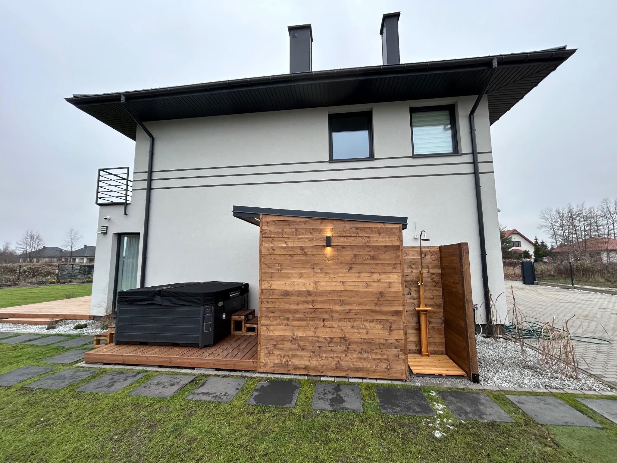 Nowoczesny taras z drewnianą zabudową sauny zewnętrznej i jacuzzi, zlokalizowany przy budynku z balkonem. Uwagę przyciąga minimalistyczny design i staranne wykonanie.