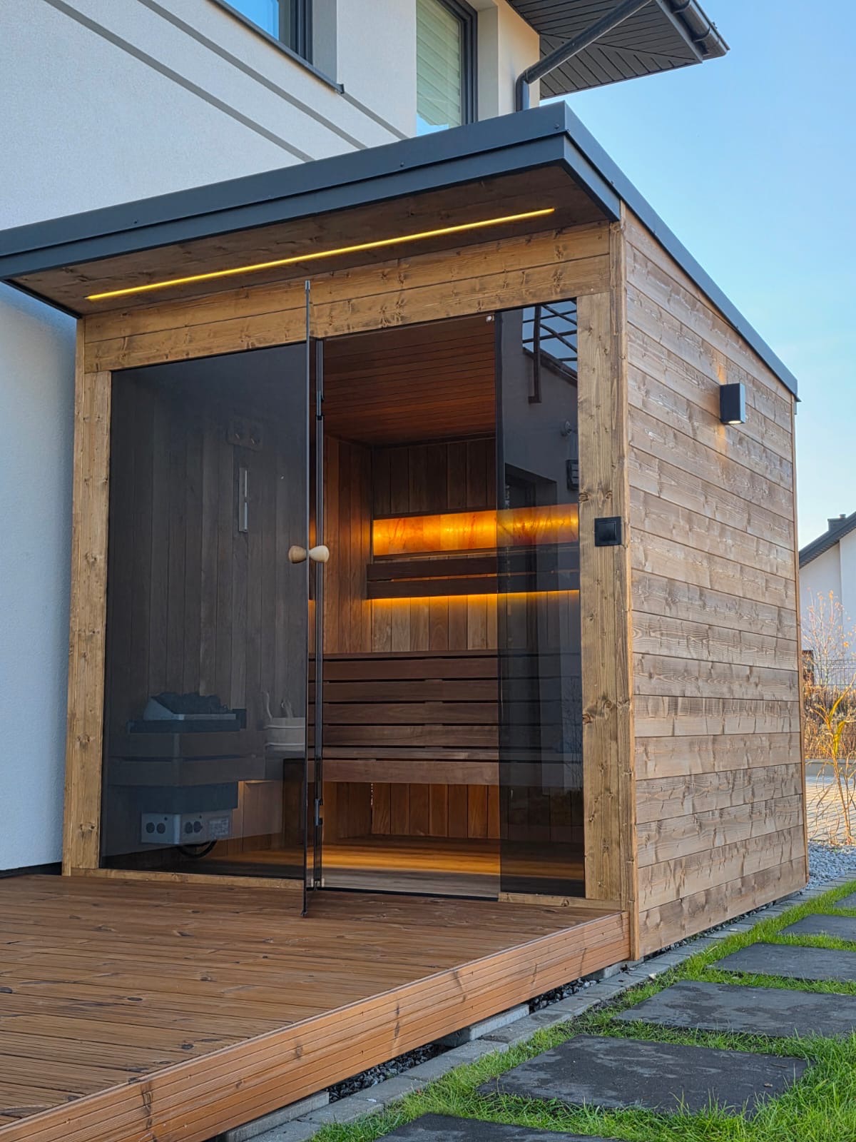 Drewniana sauna zewnętrzna z przeszklonymi drzwiami, oświetleniem LED i drewnianym tarasem, umieszczona obok domu z elementami nowoczesnej architektury.