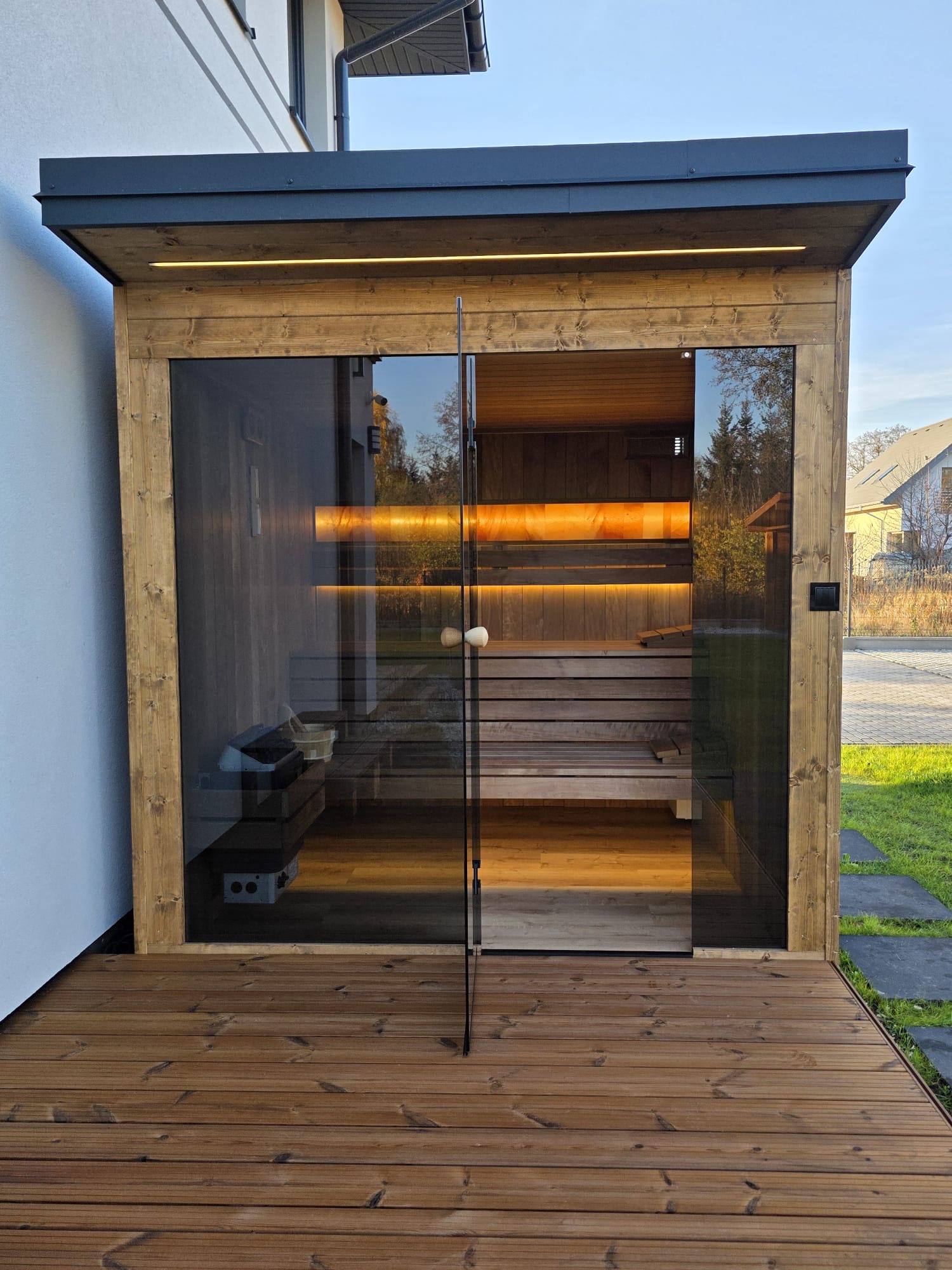 Nowoczesna sauna zewnętrzna z przeszklonymi drzwiami, oświetleniem LED i drewnianym wykończeniem, umieszczona na tarasie z desek, z widokiem na ogród.