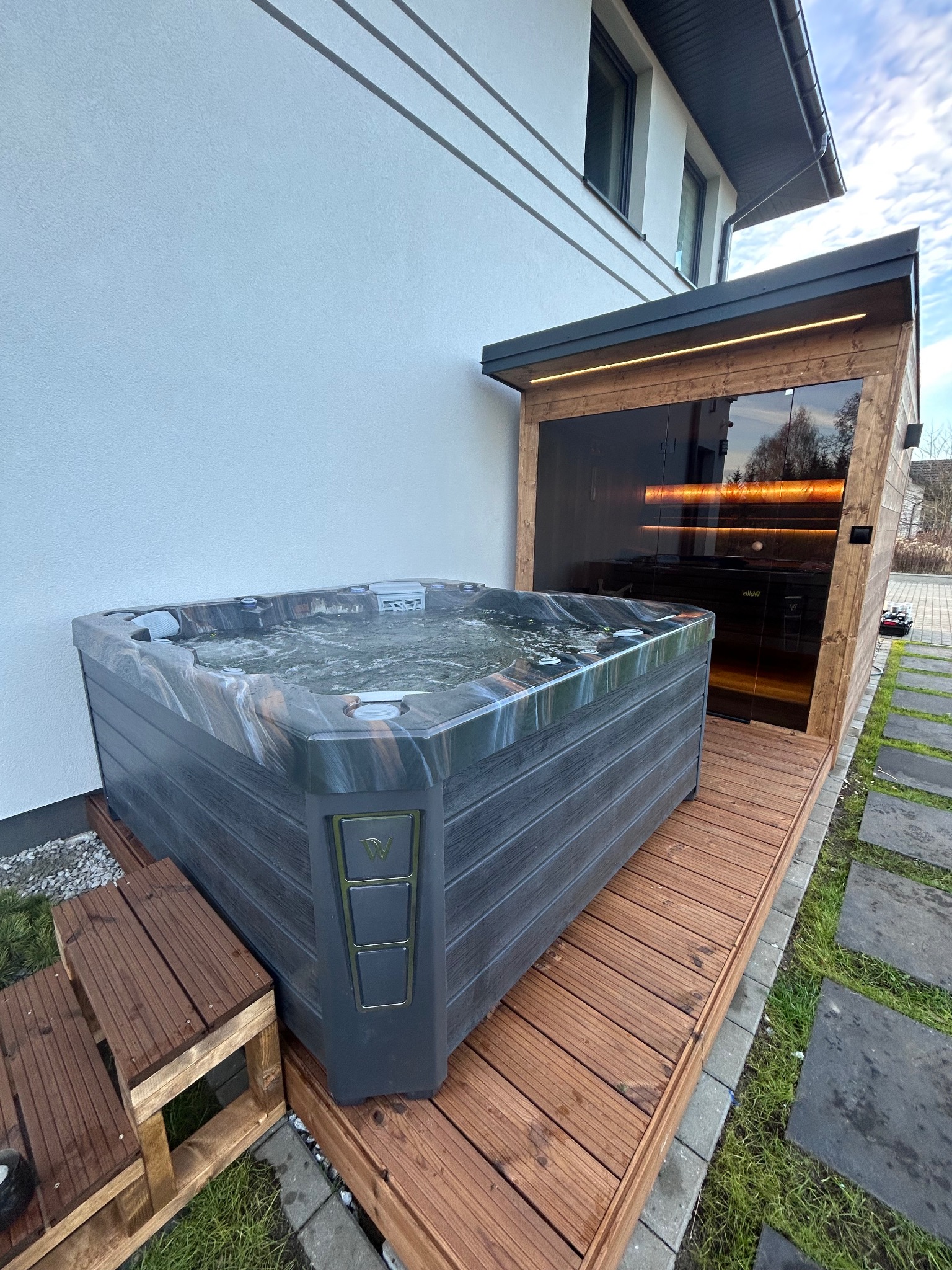 Nowoczesna sauna z przeszkloną ścianą i jacuzzi na drewnianym tarasie przy białym budynku. Oświetlenie LED podkreśla architekturę.