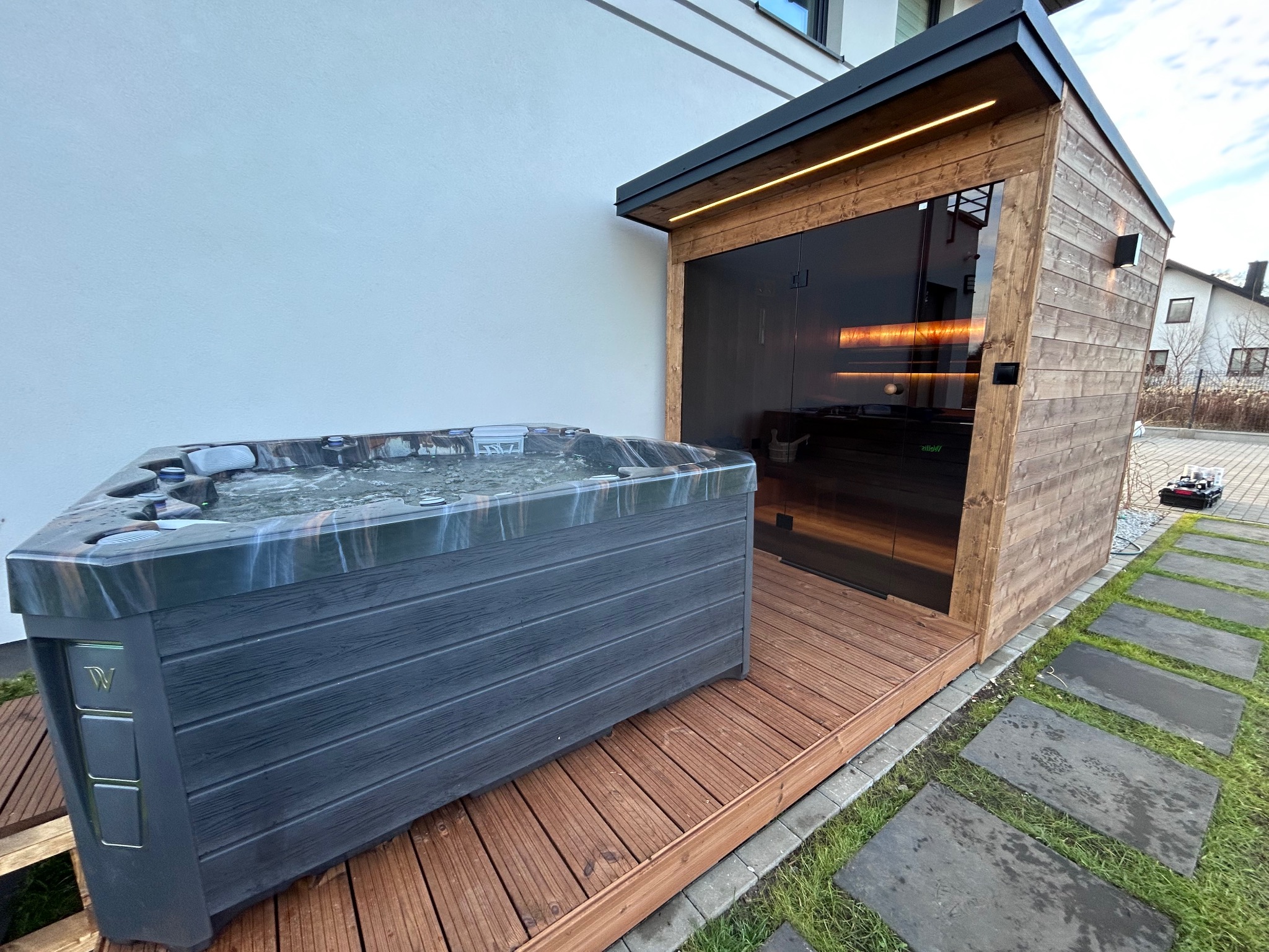 Nowoczesna sauna z przeszklonym wejściem i oświetleniem LED, obok jacuzzi na drewnianym tarasie, otoczona zielenią i kamiennymi płytami chodnikowymi.