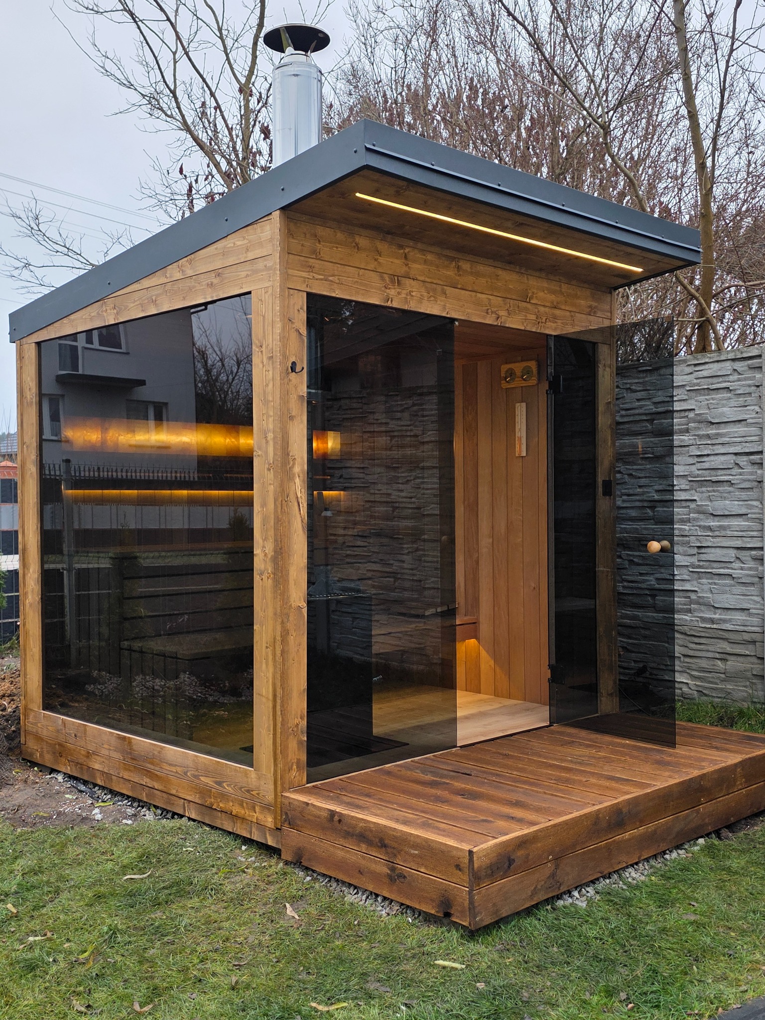 Nowoczesna sauna ogrodowa z drewnianą konstrukcją, przeszklonymi ścianami i tarasem. Widoczny komin i oświetlenie LED. Tło stanowią drzewa i ogrodzenie.
