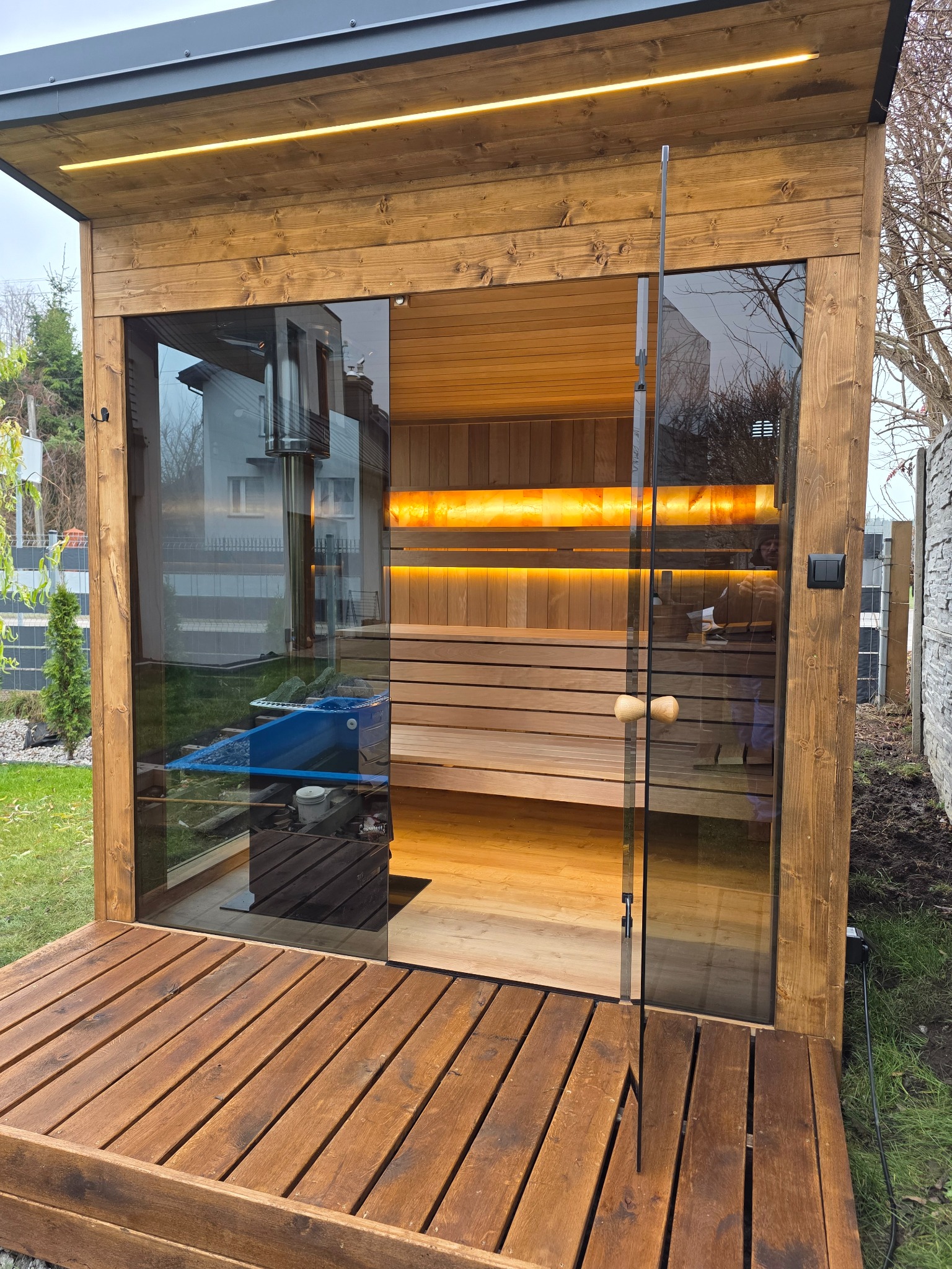 Drewniana sauna zewnętrzna z przeszklonymi drzwiami, oświetleniem LED i tarasem z desek. Widoczne wnętrze sauny z piecem i panelami solnymi.