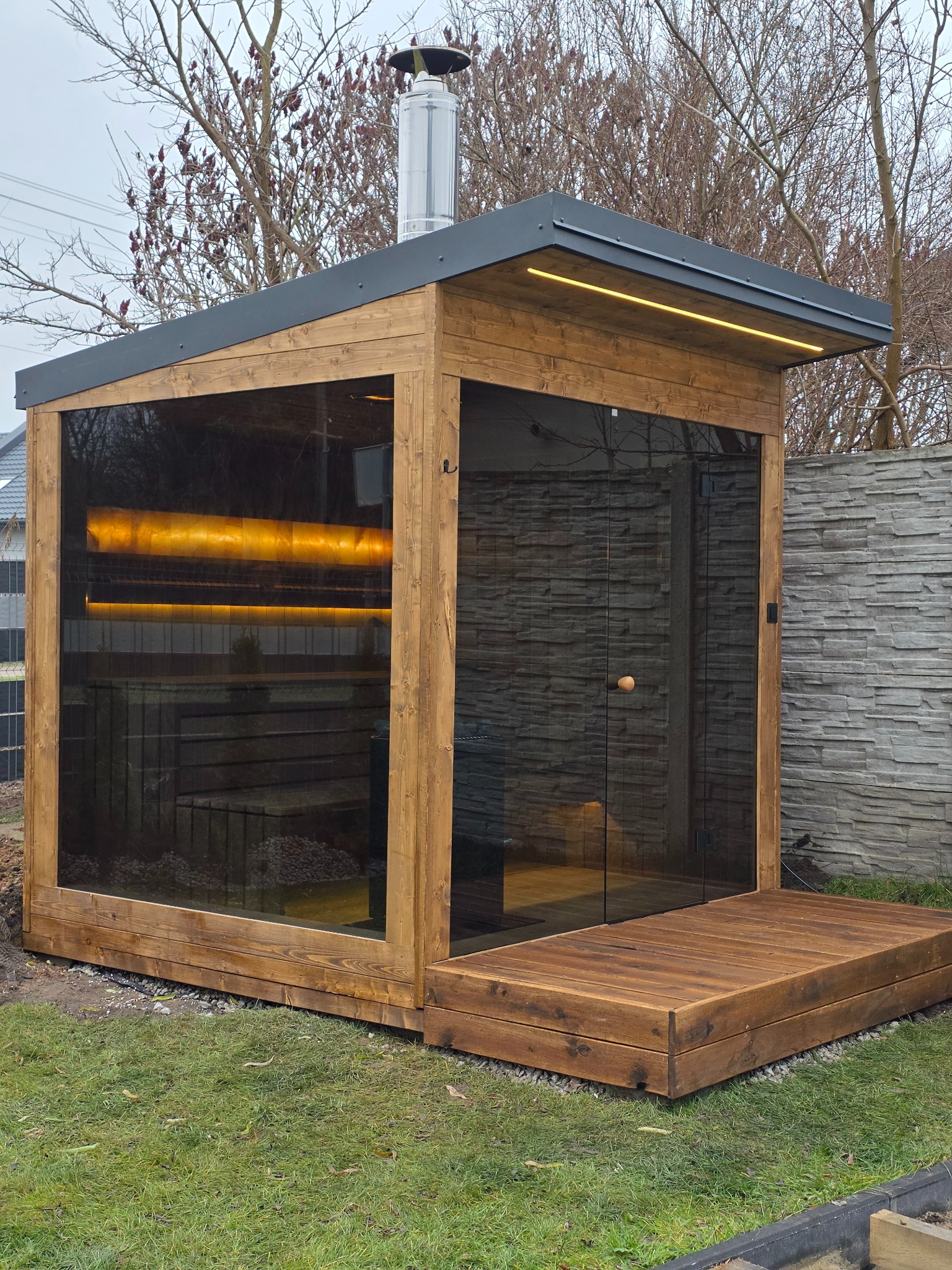 Nowoczesna sauna ogrodowa z drewna i szkła, z oświetleniem LED i kominem, na tle zielonego trawnika i kamiennego muru.