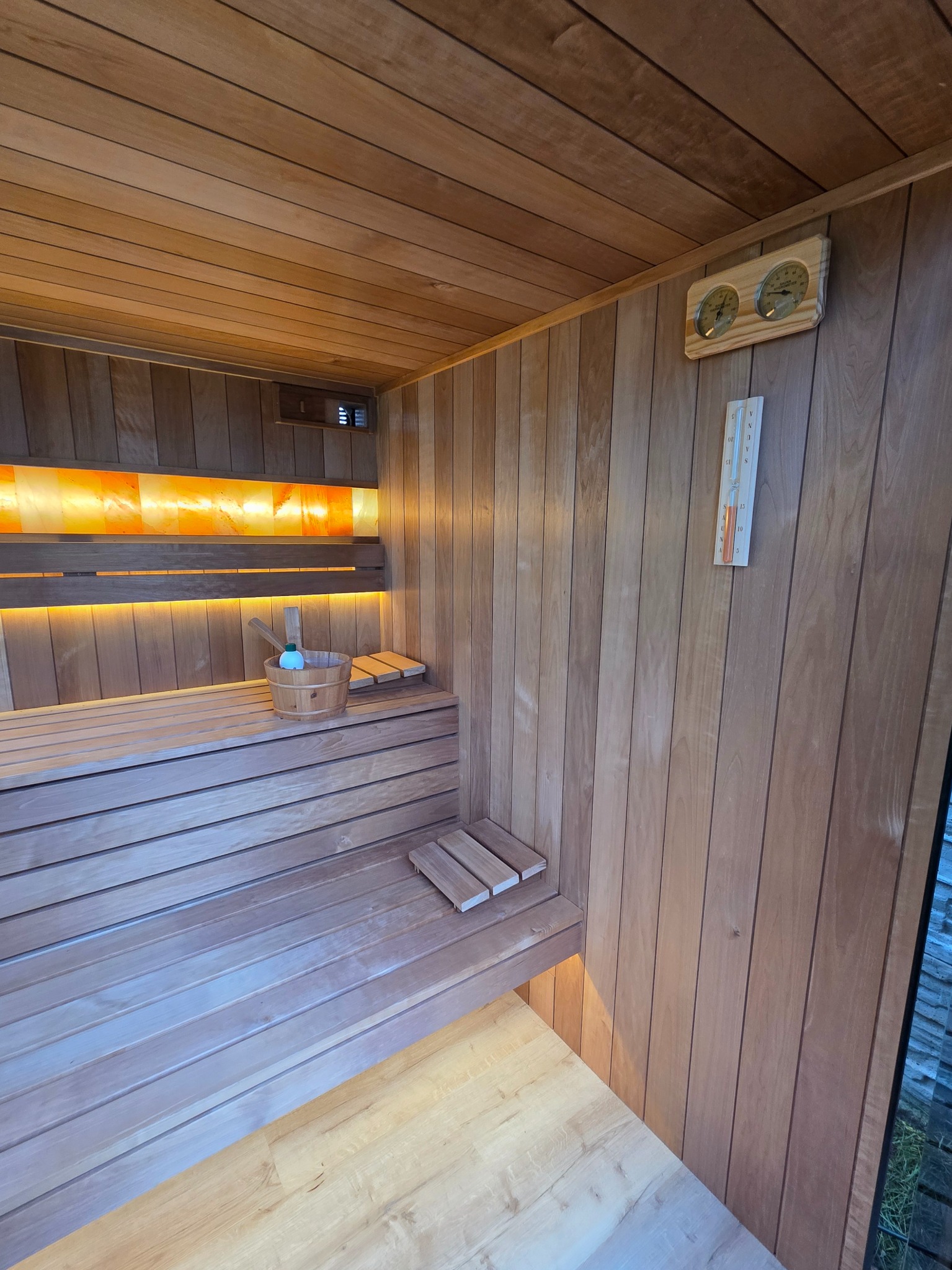 Drewniana sauna z oświetleniem solnym, termometrem i higrometrem. Wnętrze wykończone boazerią, z drewnianymi ławkami i akcesoriami do sauny.