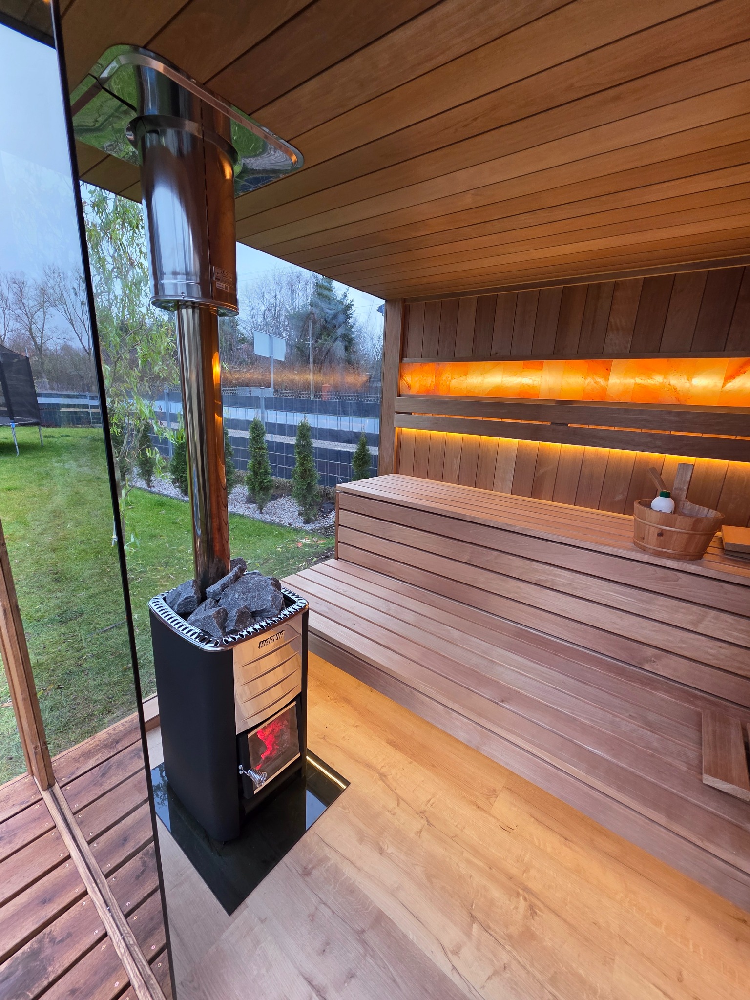 Nowoczesna sauna z piecem Harvia, drewnianymi ławkami i podświetlanymi panelami solnymi. Widok z zewnątrz przez szklaną ścianę na zielony trawnik.