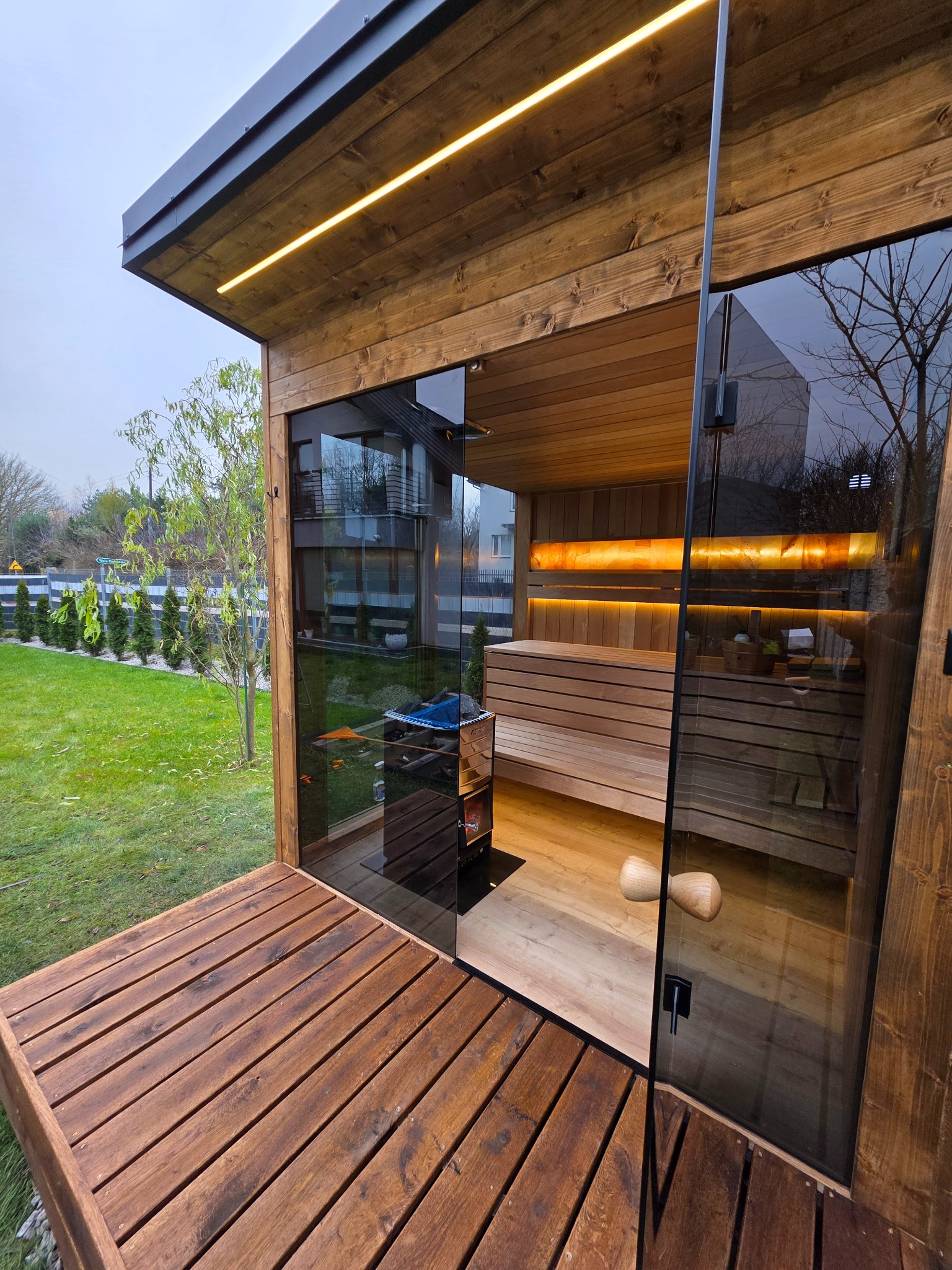 Nowoczesna sauna zewnętrzna z drewnianym tarasem i przeszklonymi ścianami, oświetlona listwami LED, z widokiem na zielony ogród i budynek mieszkalny w tle.