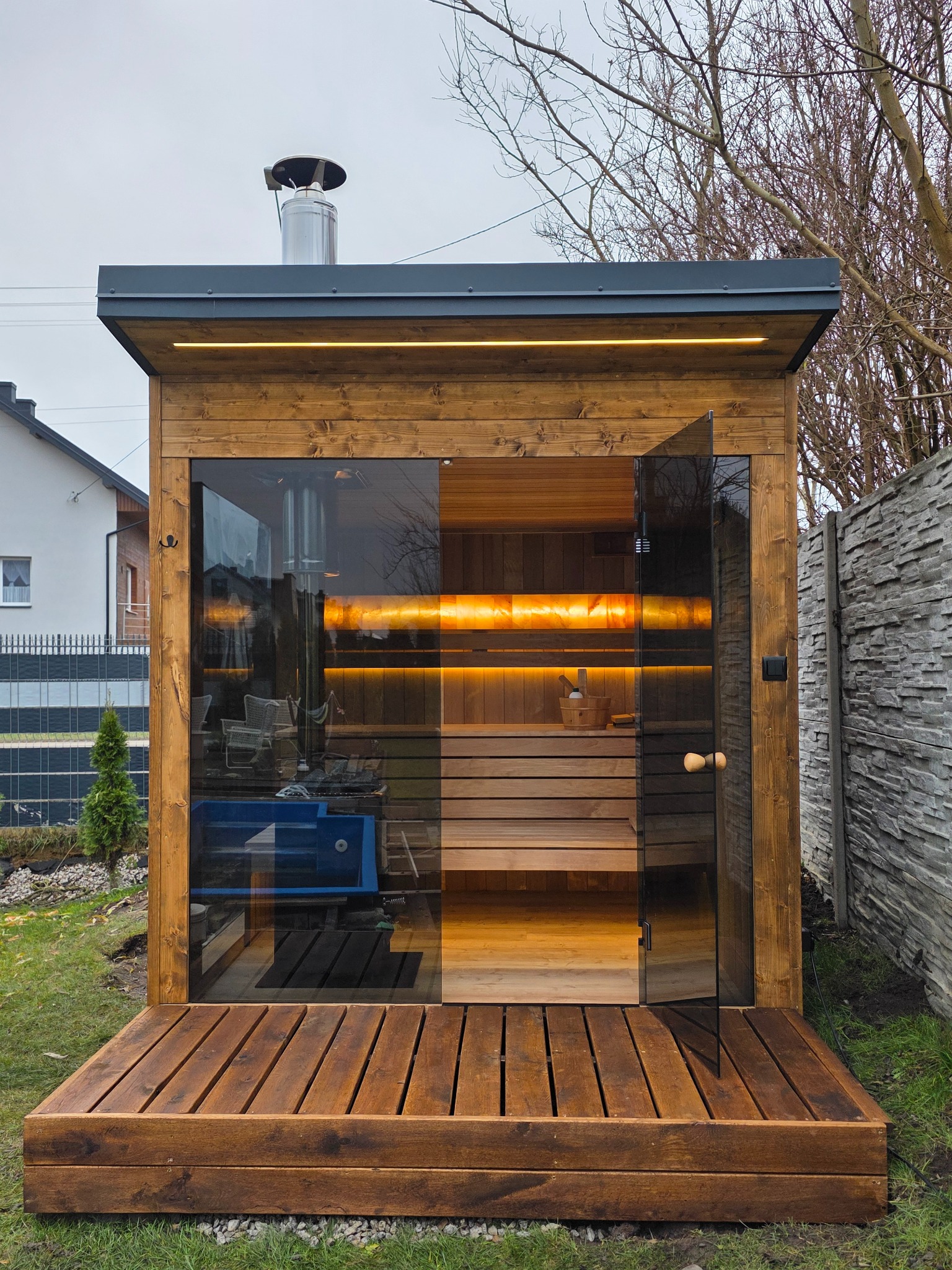 Drewniana sauna zewnętrzna z przeszklonymi drzwiami, oświetleniem LED i kominem, umieszczona na tarasie z desek. Widok na wnętrze sauny i otoczenie.