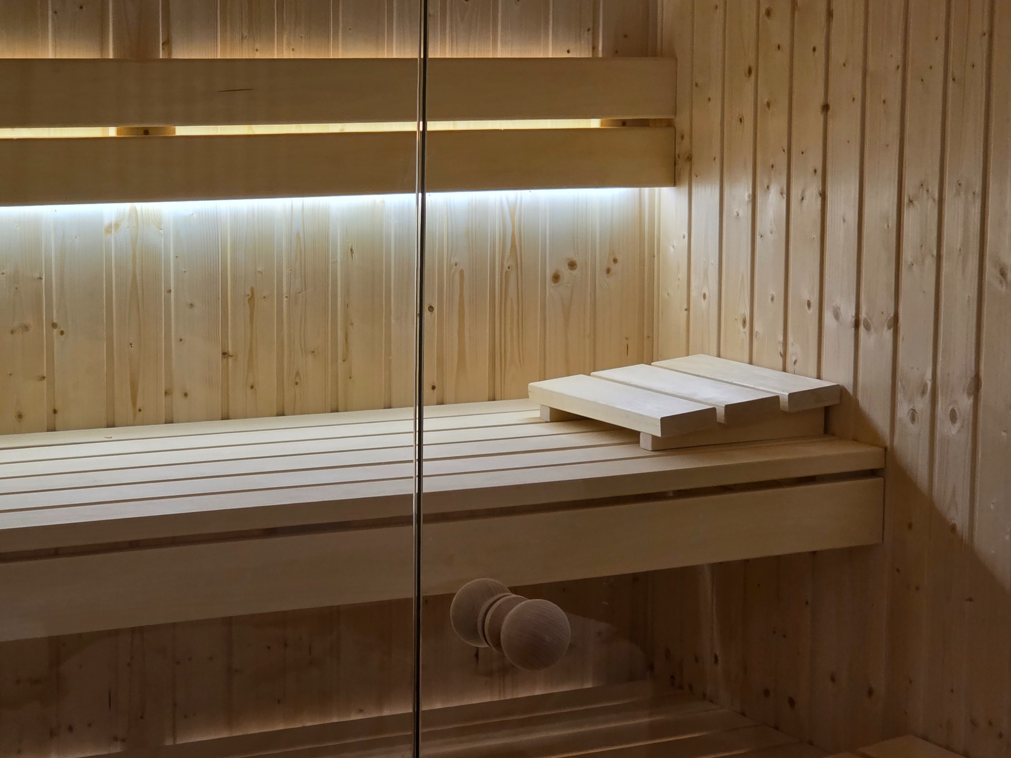 Jasne drewno i szklane drzwi w saunie z podświetleniem LED. Wnętrze sauny z ławkami i zagłówkiem. Minimalistyczny design.