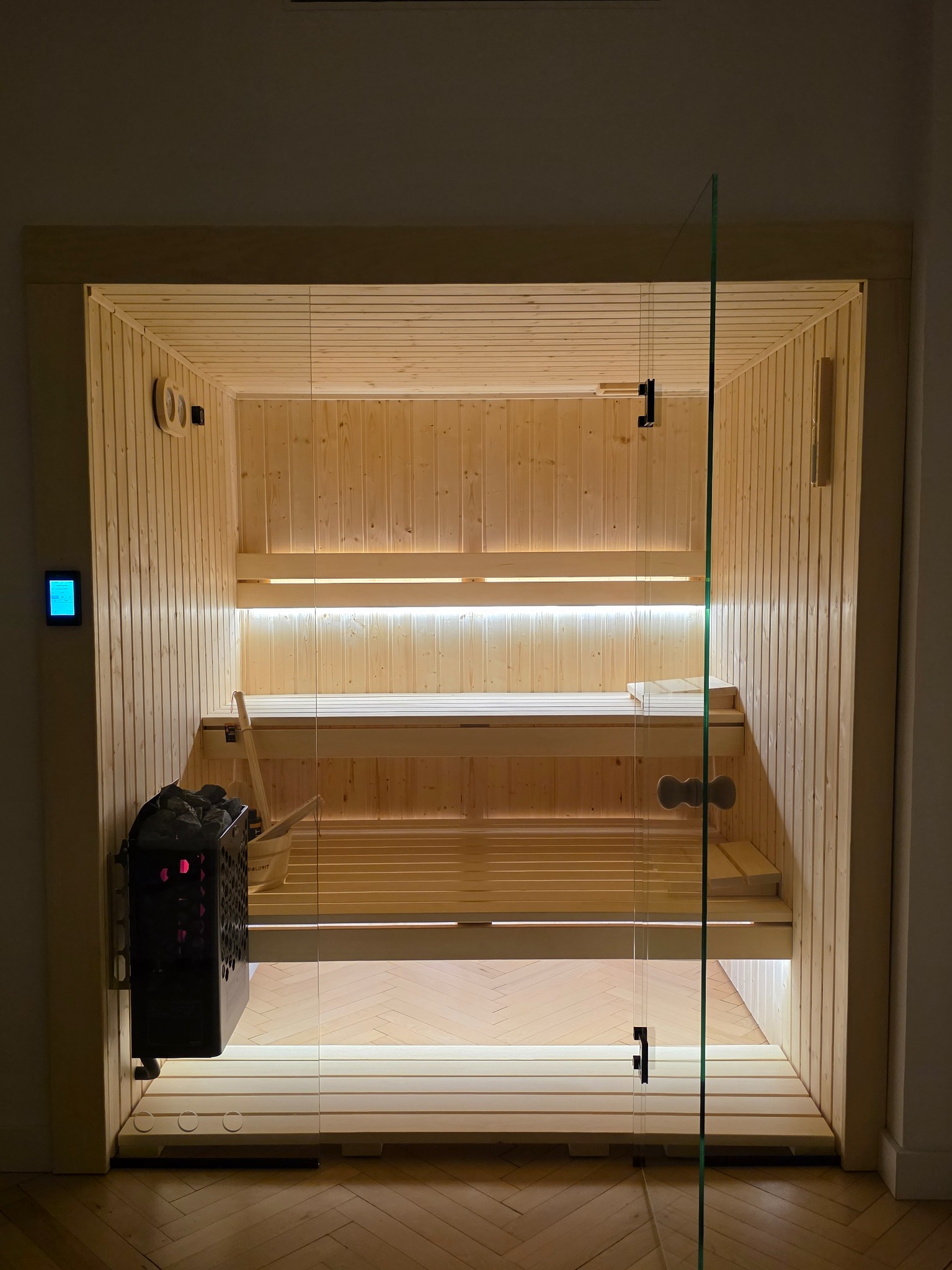 Nowoczesna sauna z przeszklonymi drzwiami, oświetleniem LED i piecem z kamieniami. Jasne drewno na ścianach i ławkach. Widoczny panel sterowania.