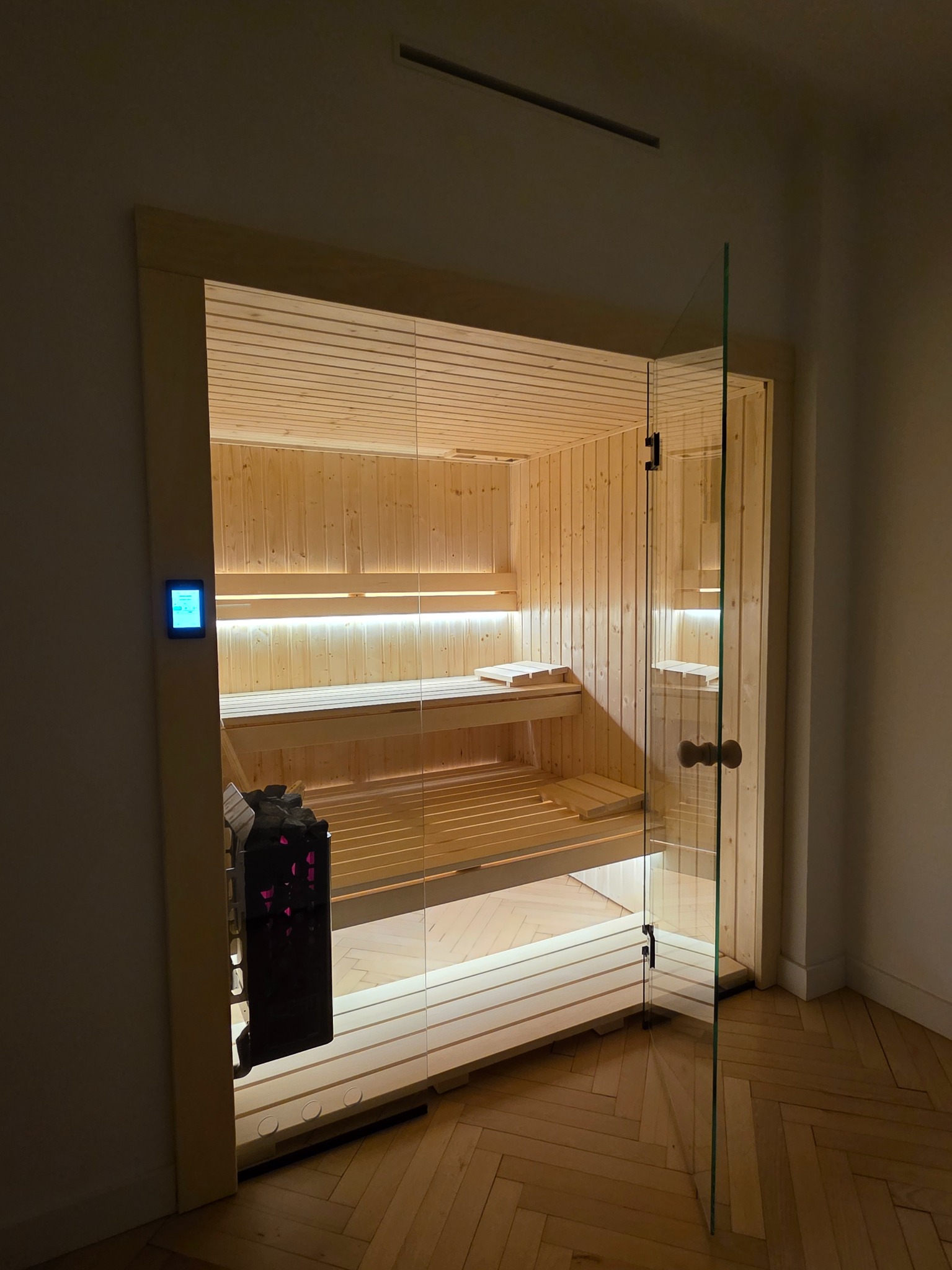 Nowoczesna sauna z przeszklonymi drzwiami, drewnianym wnętrzem i podświetleniem LED, widoczny piec z kamieniami. Parkiet na podłodze.