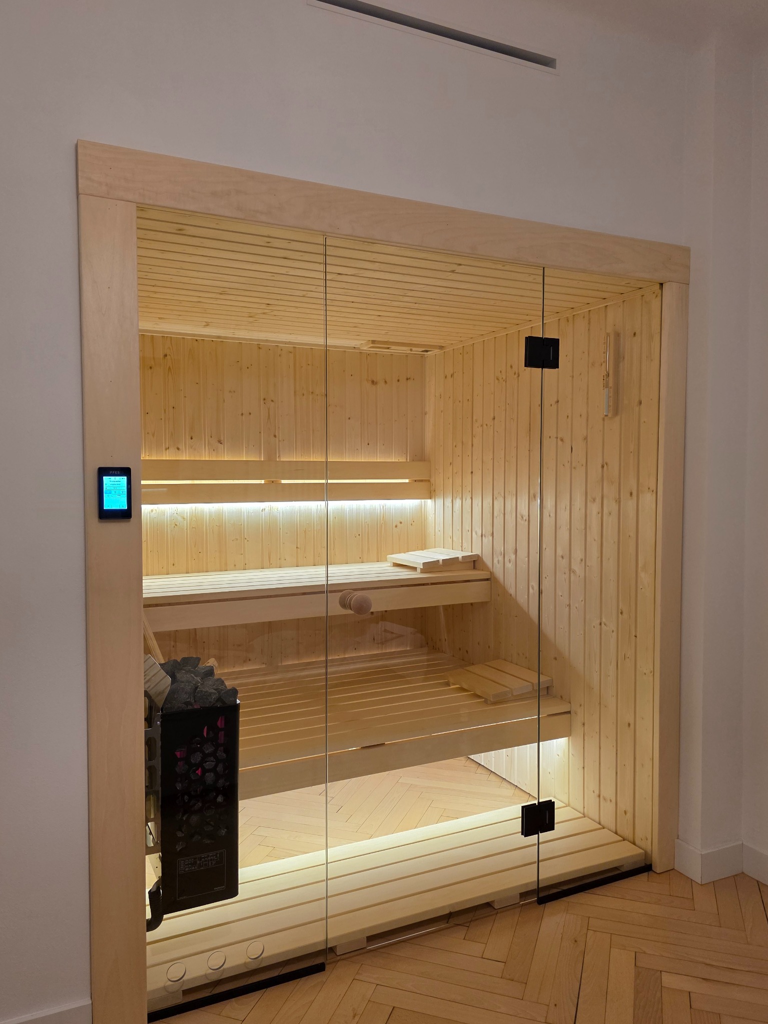 Jasna, drewniana sauna z przeszklonymi drzwiami i piecem na kamienie. Widoczne drewniane ławki i oświetlenie LED. Minimalistyczny design, parkiet na podłodze.