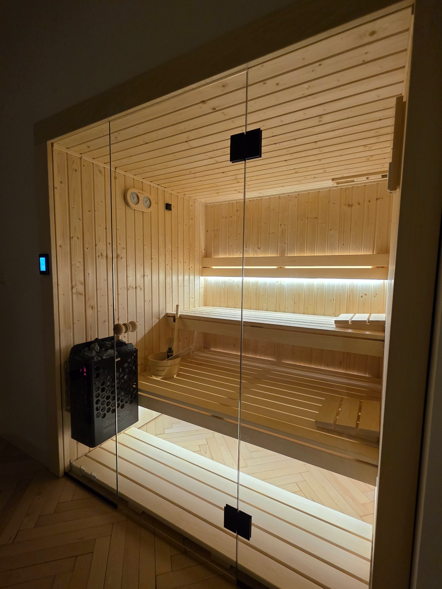 Jasne drewno i szkło w nowoczesnej saunie z piecem na kamienie, oświetleniem LED i akcesoriami. Parkiet w jodełkę na podłodze.