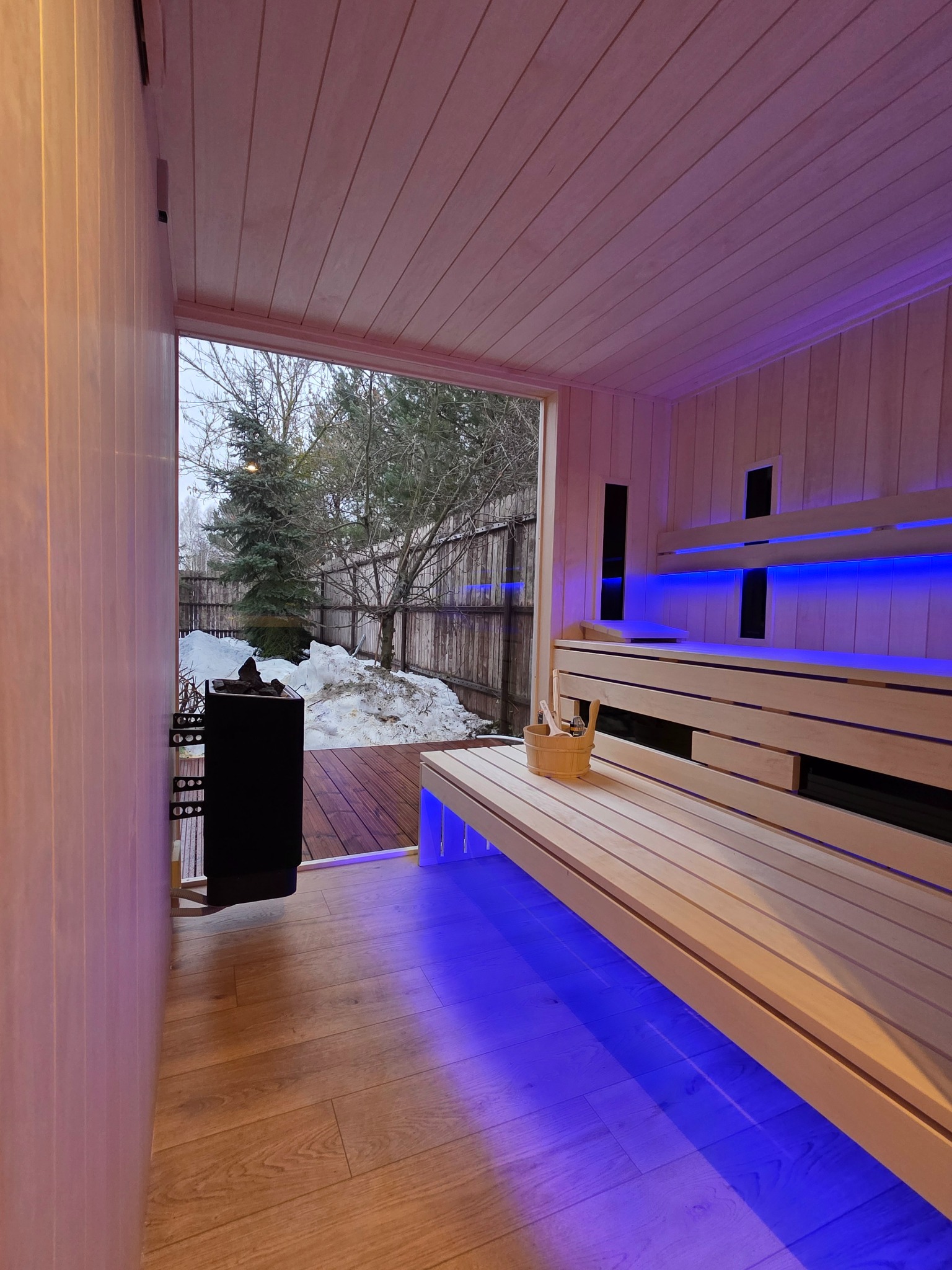 Nowoczesna sauna z drewnianym wykończeniem, podświetleniem LED i widokiem na zimowy ogród przez duże okno. Piec na kamienie i akcesoria do sauny na ławce.