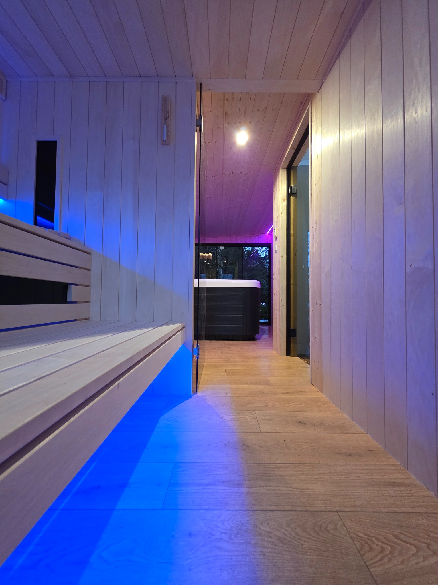 Nowoczesna sauna z drewnianymi ławkami podświetlonymi na niebiesko, widok na jacuzzi przez szklane drzwi. Drewniane wykończenie ścian i podłogi.