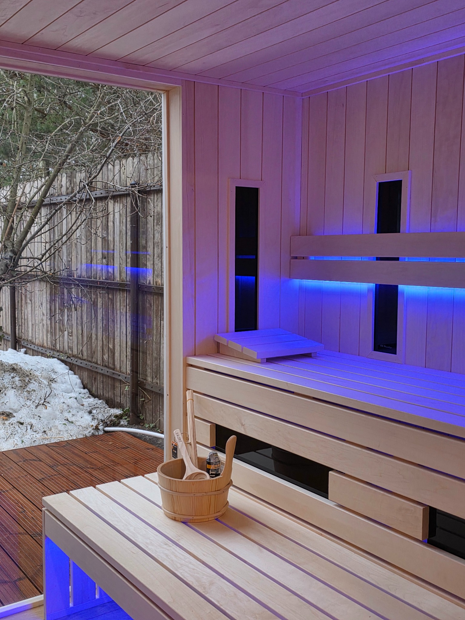 Wnętrze sauny z drewnianym wykończeniem i niebieskim oświetleniem LED. Na pierwszym planie wiadro z akcesoriami, za oknem widok na zimowy ogród.