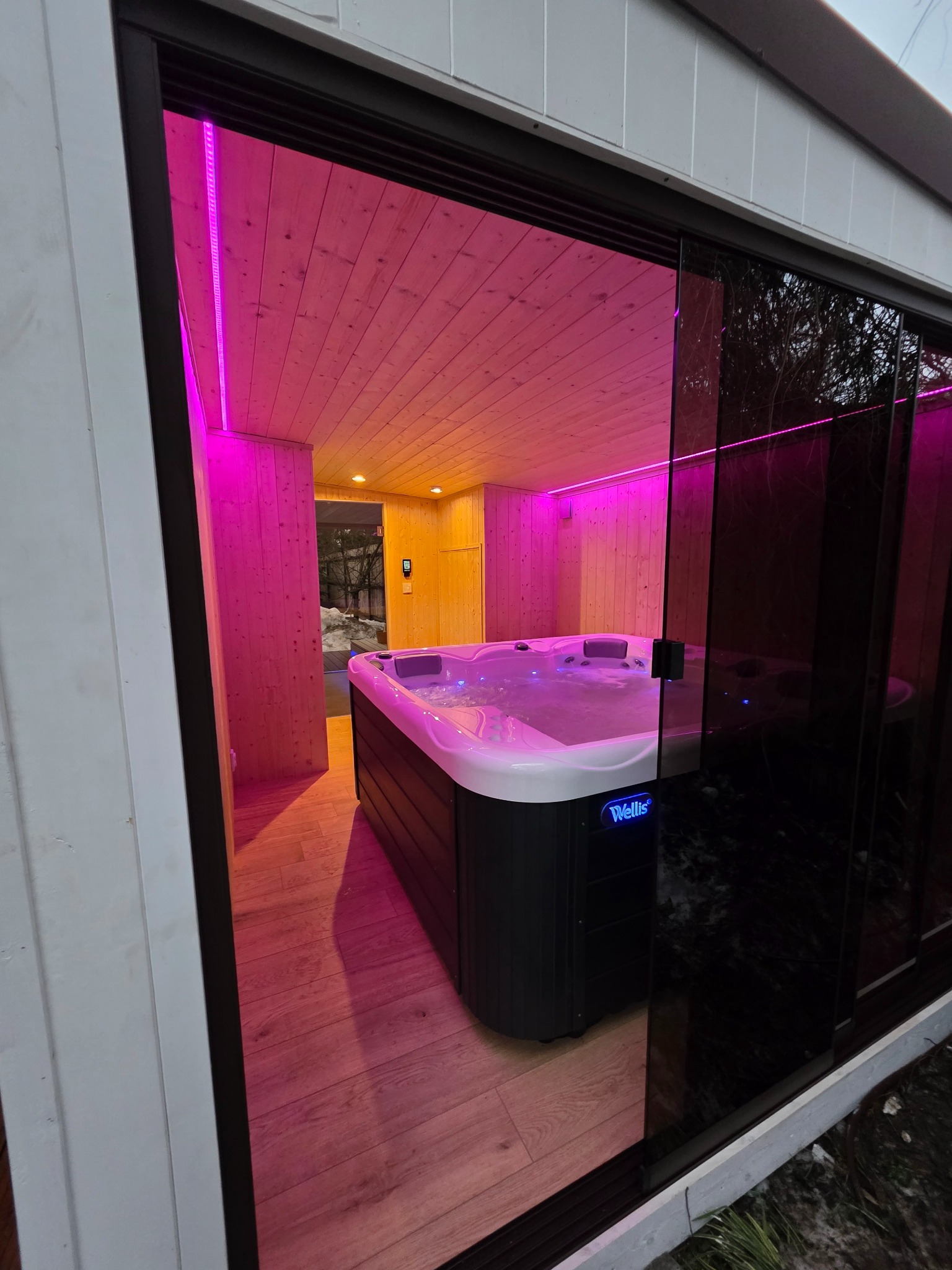 Nowoczesna sauna z jacuzzi, oświetlona różowym światłem LED, widoczna przez przesuwne szklane drzwi. Drewniane wykończenie wnętrza i podłogi.