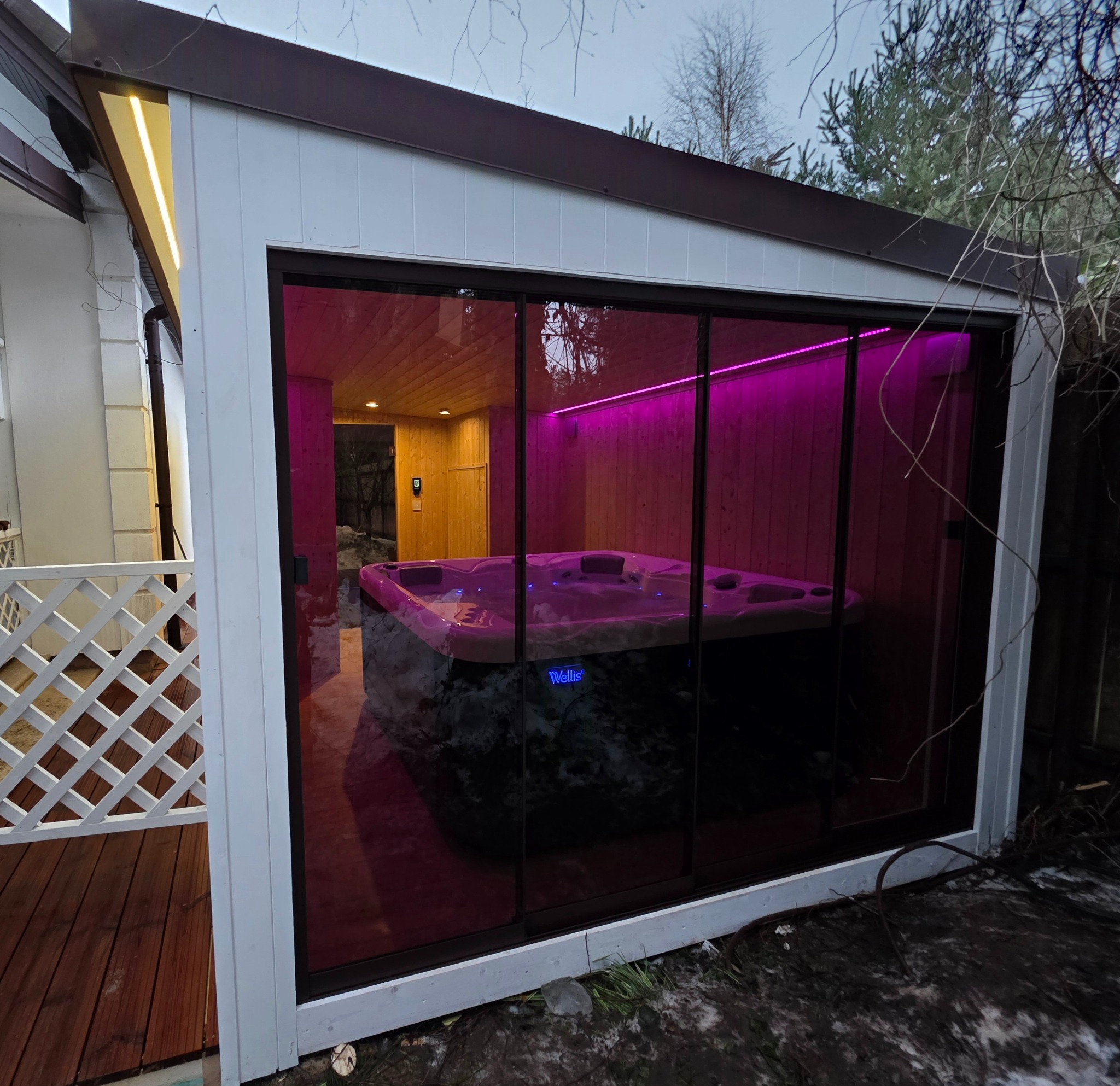 Nowoczesna sauna zewnętrzna z jacuzzi, oświetlona fioletowym światłem LED, widoczna przez szklane drzwi przesuwne. Obok drewniany taras z białą balustradą.