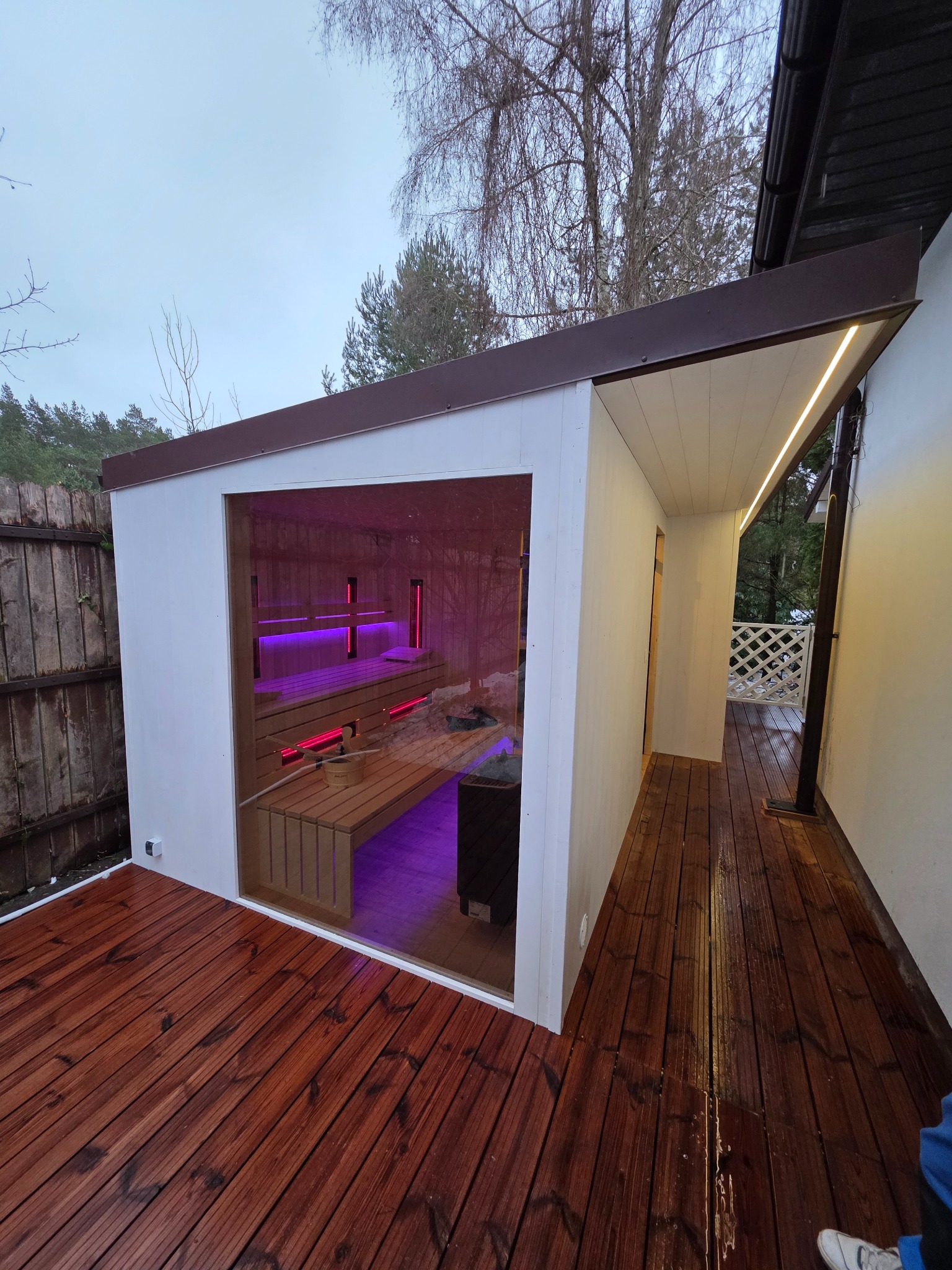 Nowoczesna sauna zewnętrzna z drewnianym tarasem i oświetleniem LED, widoczna przez duże okno. Wnętrze sauny podświetlone na różowo.