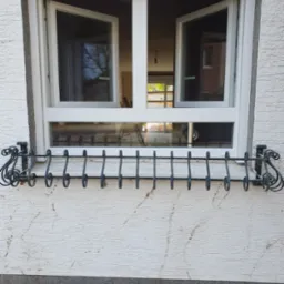 Białe okno PCV z otwartymi skrzydłami, ozdobny, metalowy kwietnik pod oknem, elewacja budynku o fakturze baranka. Widoczny fragment wnętrza z drabiną.