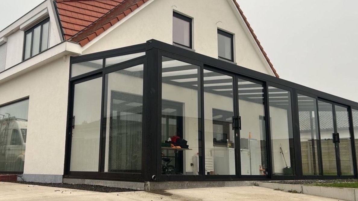 Nowoczesna, przeszklona zabudowa tarasu z czarnymi ramami, przylegająca do białego domu z czerwonym dachem. Widoczny fragment zaparkowanego busa.