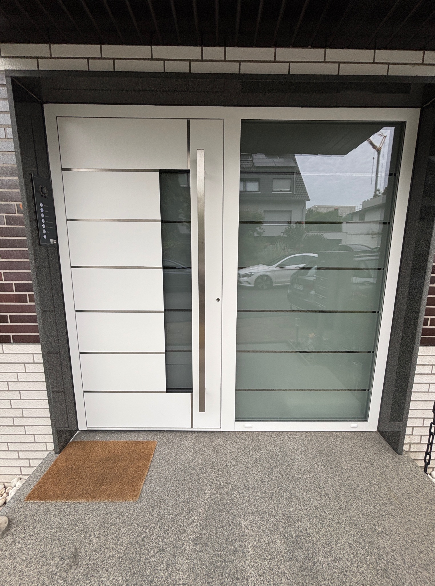 Nowoczesne, białe drzwi wejściowe z przeszkleniem, z długim, pionowym uchwytem, zamontowane w elewacji z cegły. Widoczny wycieraczka i fragment domu w odbiciu szyby.