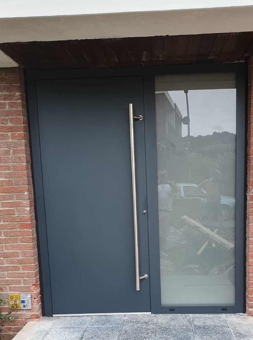 Grafitowe drzwi zewnętrzne z długim, metalowym uchwytem i szybą, wkomponowane w ceglaną elewację domu. W szybie odbijają się postacie i samochód.