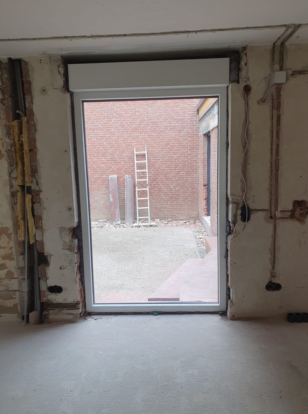 Białe okno z roletą zewnętrzną w surowym otworze ściennym. Widok na ceglany mur i drabinę. Prace wykończeniowe, w tle podwórko z płytami.