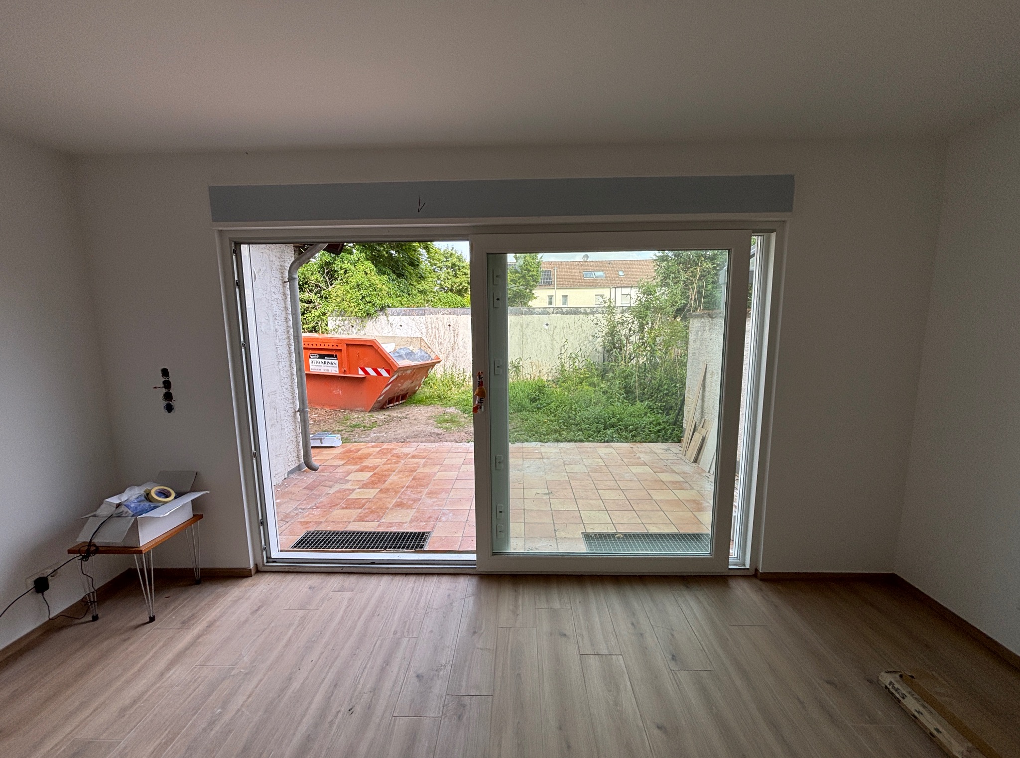 Wnętrze pokoju z dużymi przesuwnymi drzwiami balkonowymi, widok na taras z pomarańczowym kontenerem na gruz w tle. Podłoga wyłożona panelami.