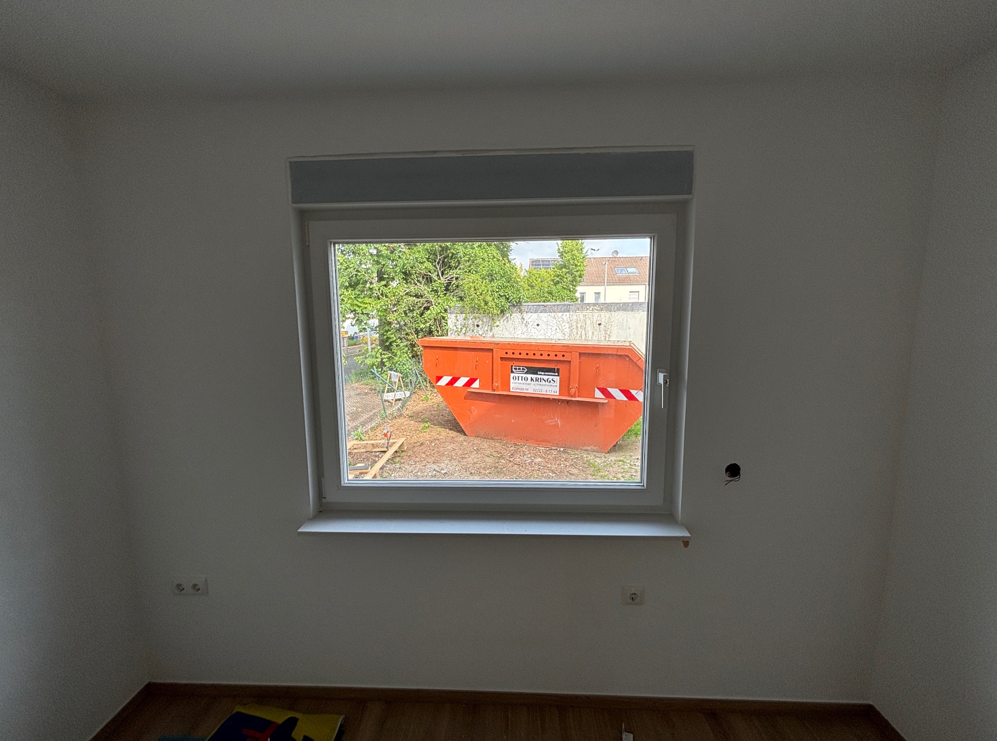 Nowe okno PCV w jasnym pokoju, widok na pomarańczowy kontener budowlany na zewnątrz. Świeżo pomalowane ściany, gniazdka elektryczne, drewniana podłoga.