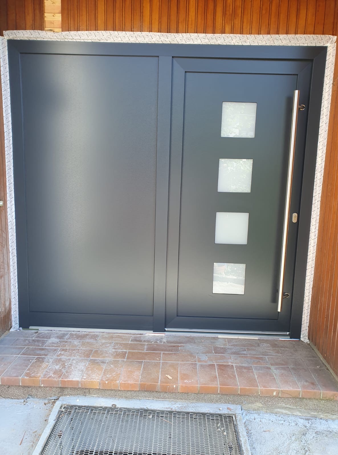 Nowoczesne, grafitowe drzwi wejściowe z czterema kwadratowymi przeszkleniami i długim, pionowym uchwytem, zamontowane w brązowej elewacji drewnianej.