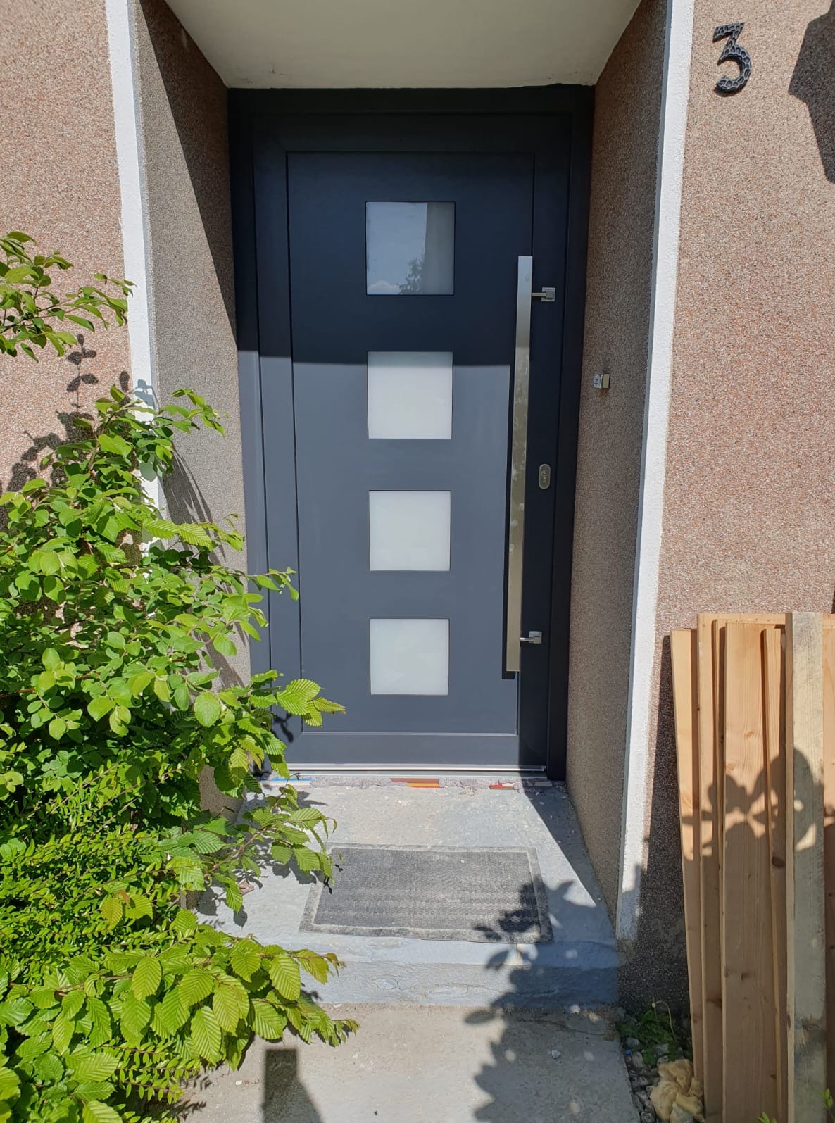 Szare drzwi zewnętrzne z trzema kwadratowymi okienkami i długim uchwytem, numer domu '3' widoczny na elewacji, otoczone zielenią i elementami budowlanymi.