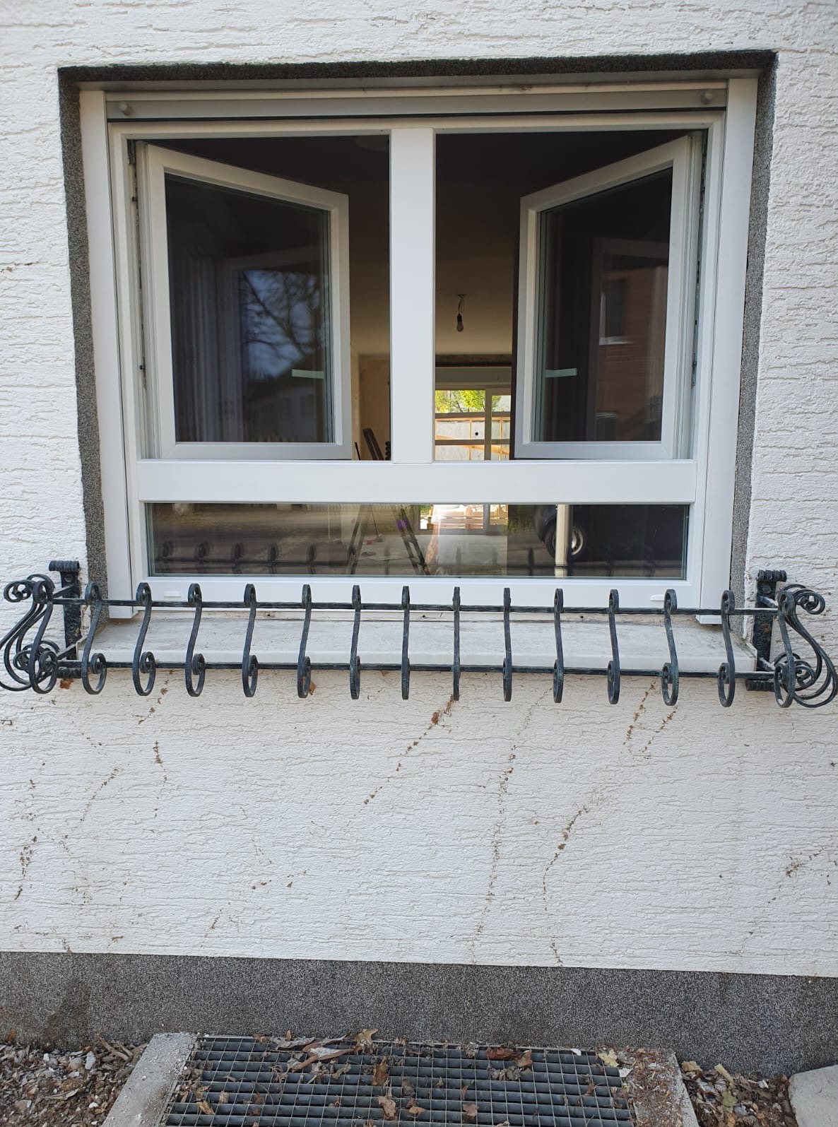 Białe okno PCV z otwartymi skrzydłami, ozdobny, metalowy kwietnik pod oknem, elewacja budynku o fakturze baranka. Widoczny fragment wnętrza z drabiną.