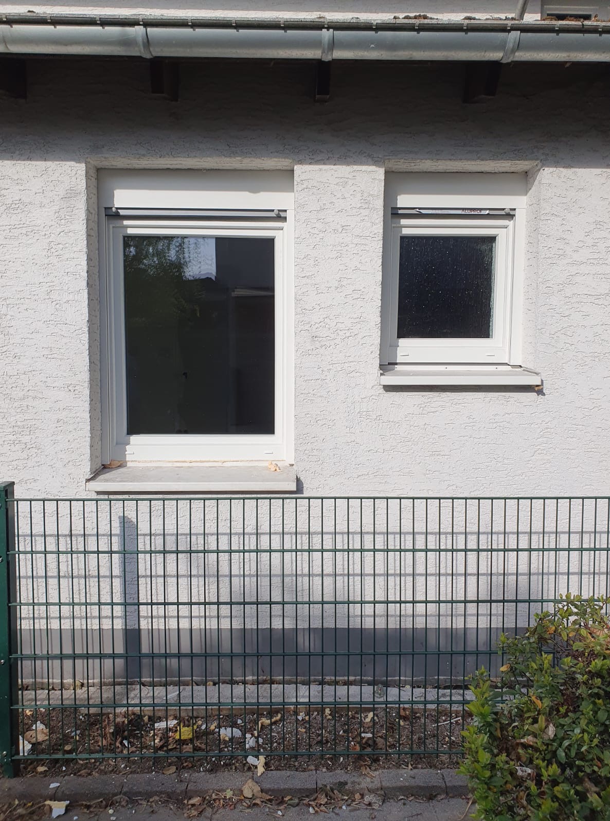 Dwa białe okna PCV o różnych rozmiarach w białej, teksturowanej ścianie budynku, widoczne zza zielonego ogrodzenia. Okno po prawej ma roletę zewnętrzną.