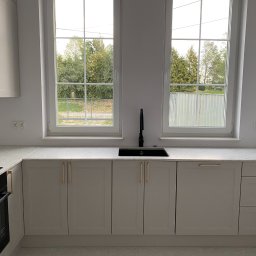 PHU Meblex II Dariusz Sawicki - Jasna kuchnia z dwoma oknami. Szare szafki z złotymi uchwytami, czarny zlew i bateria. Blat z jasnego marmuru. Widok na zieleń za oknem.