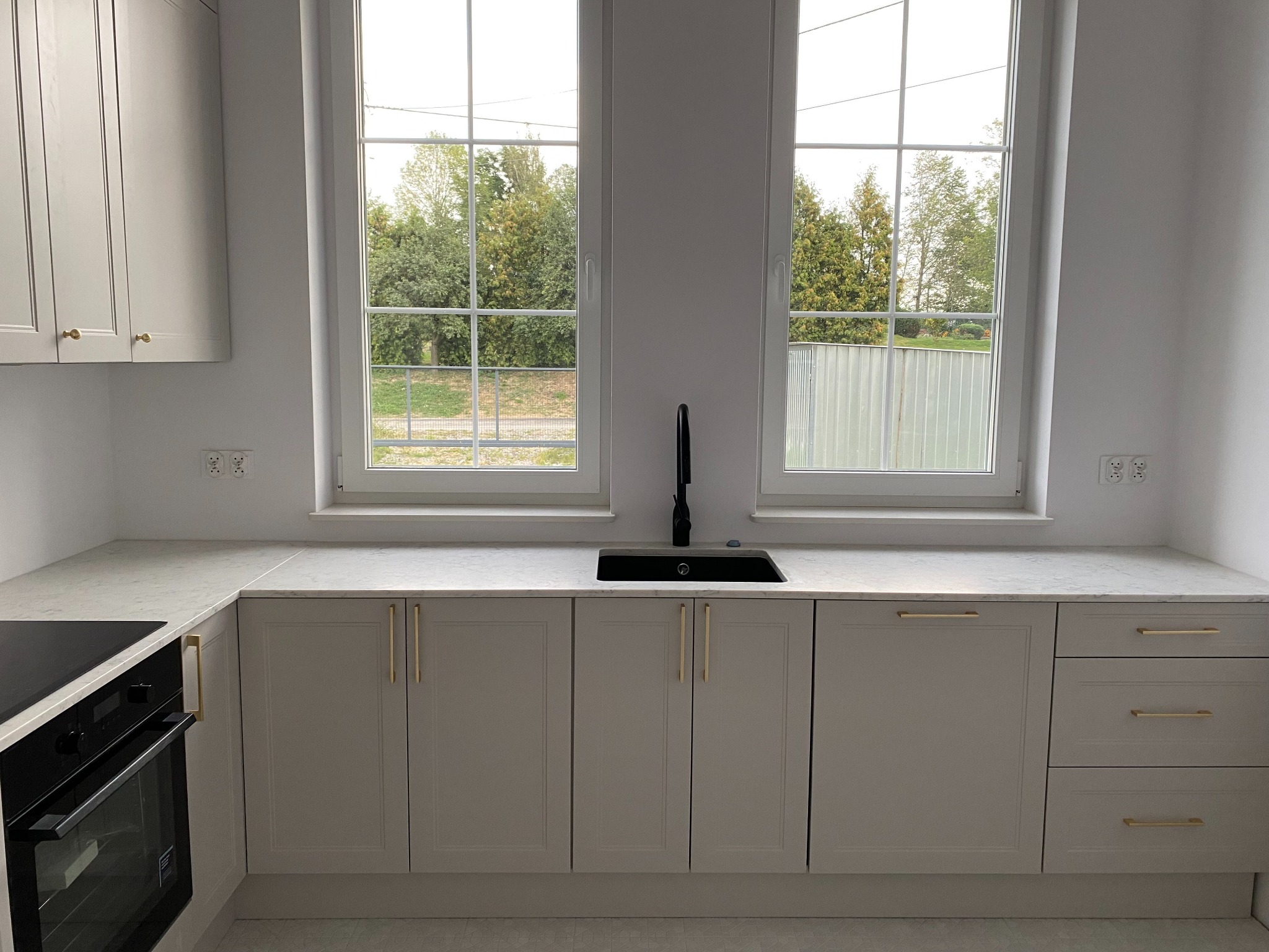 Jasna kuchnia z dwoma oknami. Szare szafki z złotymi uchwytami, czarny zlew i bateria. Blat z jasnego marmuru. Widok na zieleń za oknem.