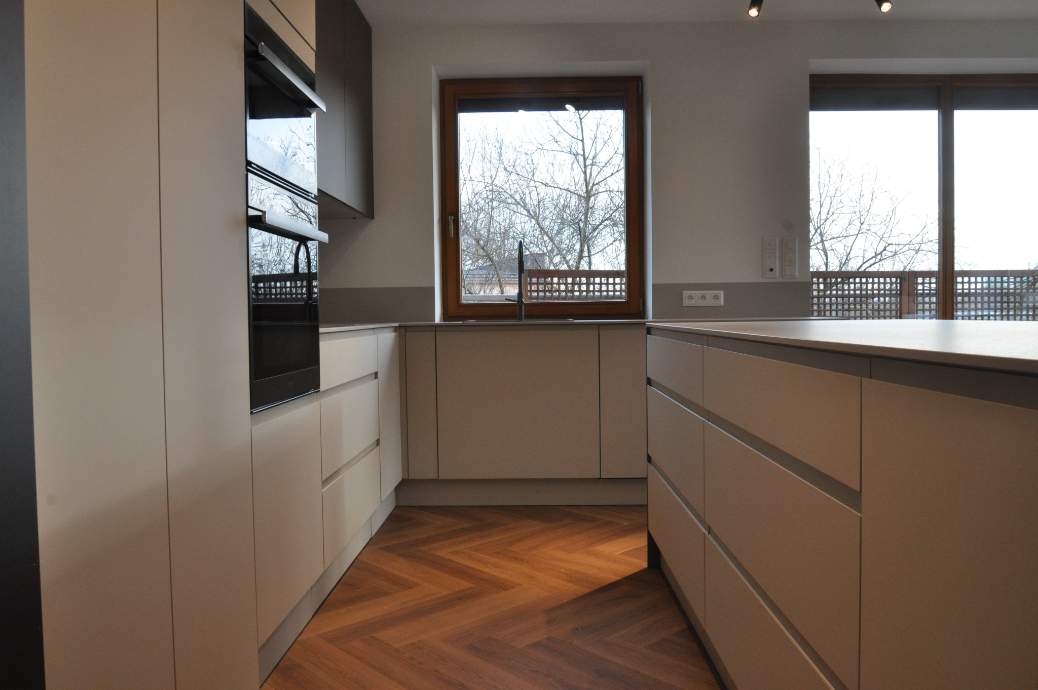 Jasna kuchnia z drewnianą podłogą w jodełkę. Widok na okno z widokiem na drzewo. Minimalistyczny design, fronty szafek bez uchwytów, nowoczesne oświetlenie.