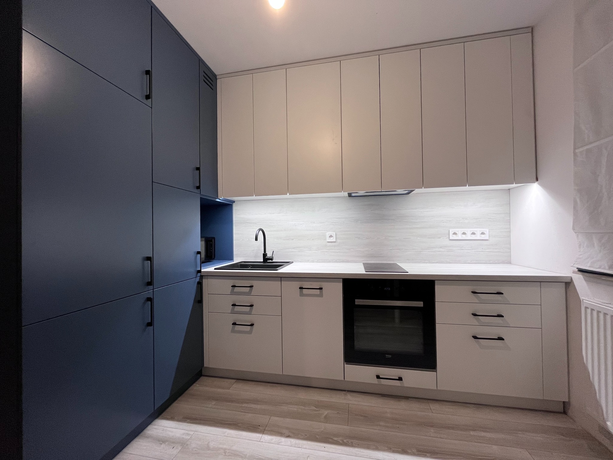 Nowoczesna, dwukolorowa kuchnia z zabudową: grafitowe szafki cargo i beżowe szafki wiszące. Czarny zlew i płyta indukcyjna na blacie. Minimalistyczny design, jasne podłogi.