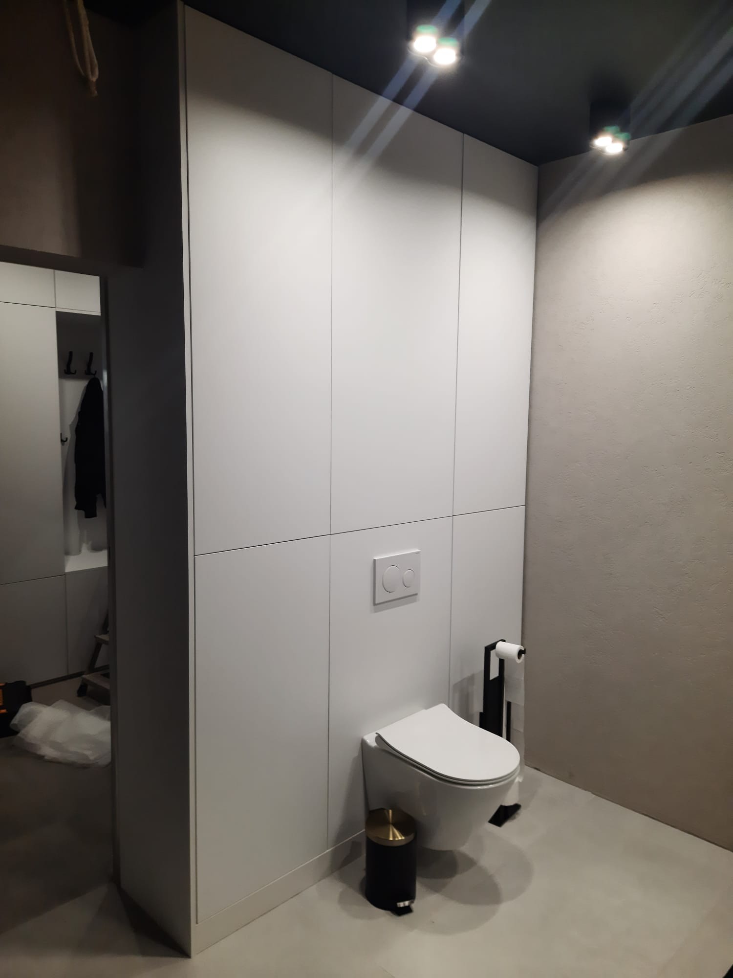 Minimalistyczna toaleta z zabudową meblową w kolorze białym, podwieszana miska WC, czarny kosz na śmieci z akcentem złota i szara ściana.